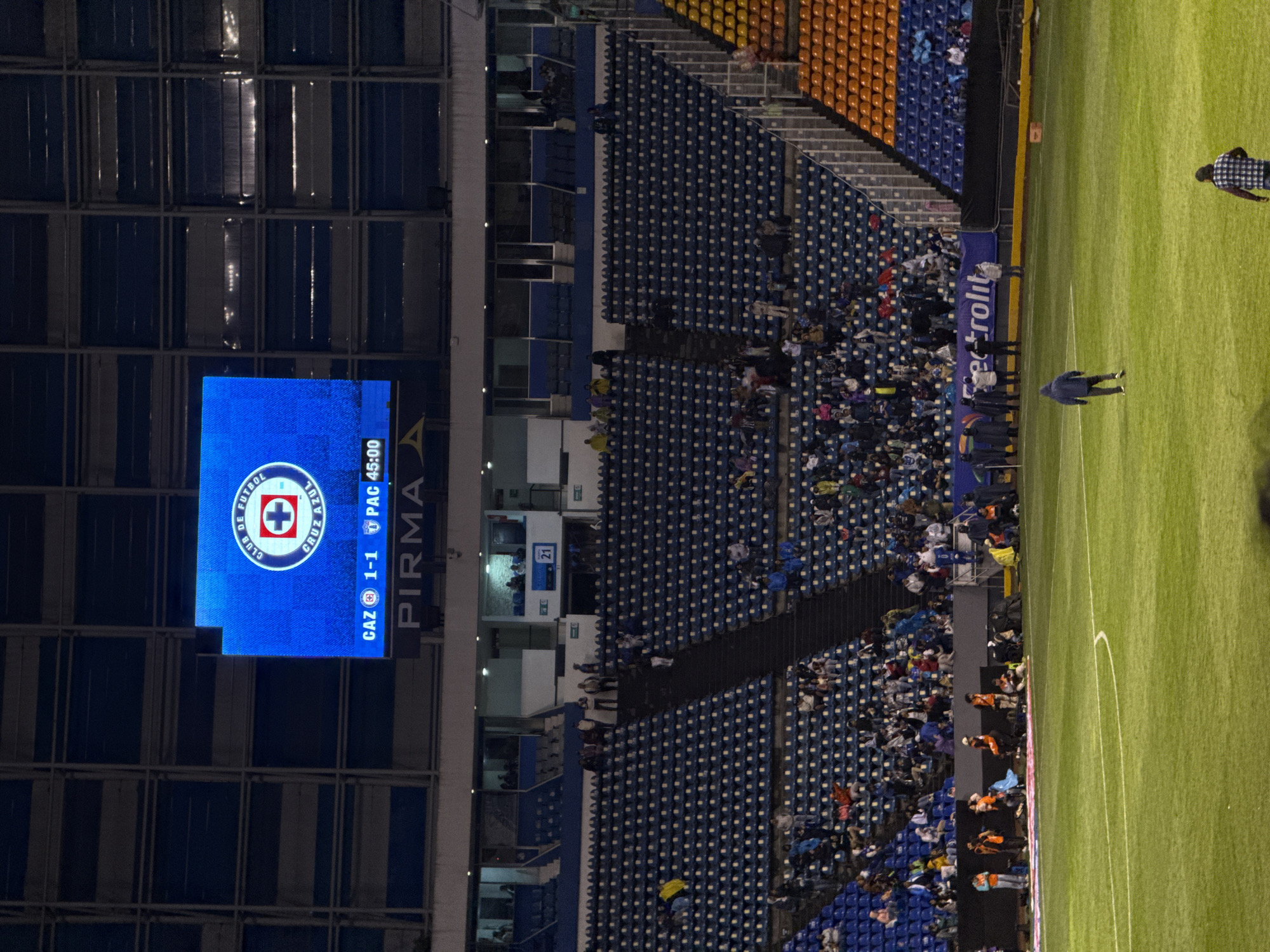 The image depicts a soccer stadium during a Cruz Azul vs. Pachuca game, likely in Puebla, Mexico. The score is tied 1-1 at the 45th minute. A large electronic scoreboard displays the Cruz Azul logo. Sections of blue and orange seats are visible, occupied by spectators. The soccer field is green, with a few people walking on it. Some staff are seen at the border of the field.