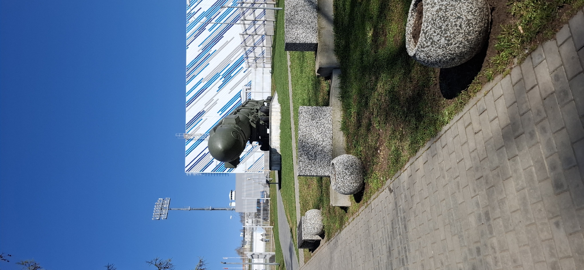 The image shows an outdoor scene in Votkinsk, Russia, featuring a monument to a ballistic missile near the Votkinsk Izhstal stadium. The monument, an olive-green missile, is positioned near a modern building with a white facade and blue and gray diagonal stripes. A green lawn separates the monument and the building from a paved walkway. Along the walkway are benches with gray stone seats and speckled gray planters. A blue sky is visible above the building and missile, and a stadium light structure stands in the background.