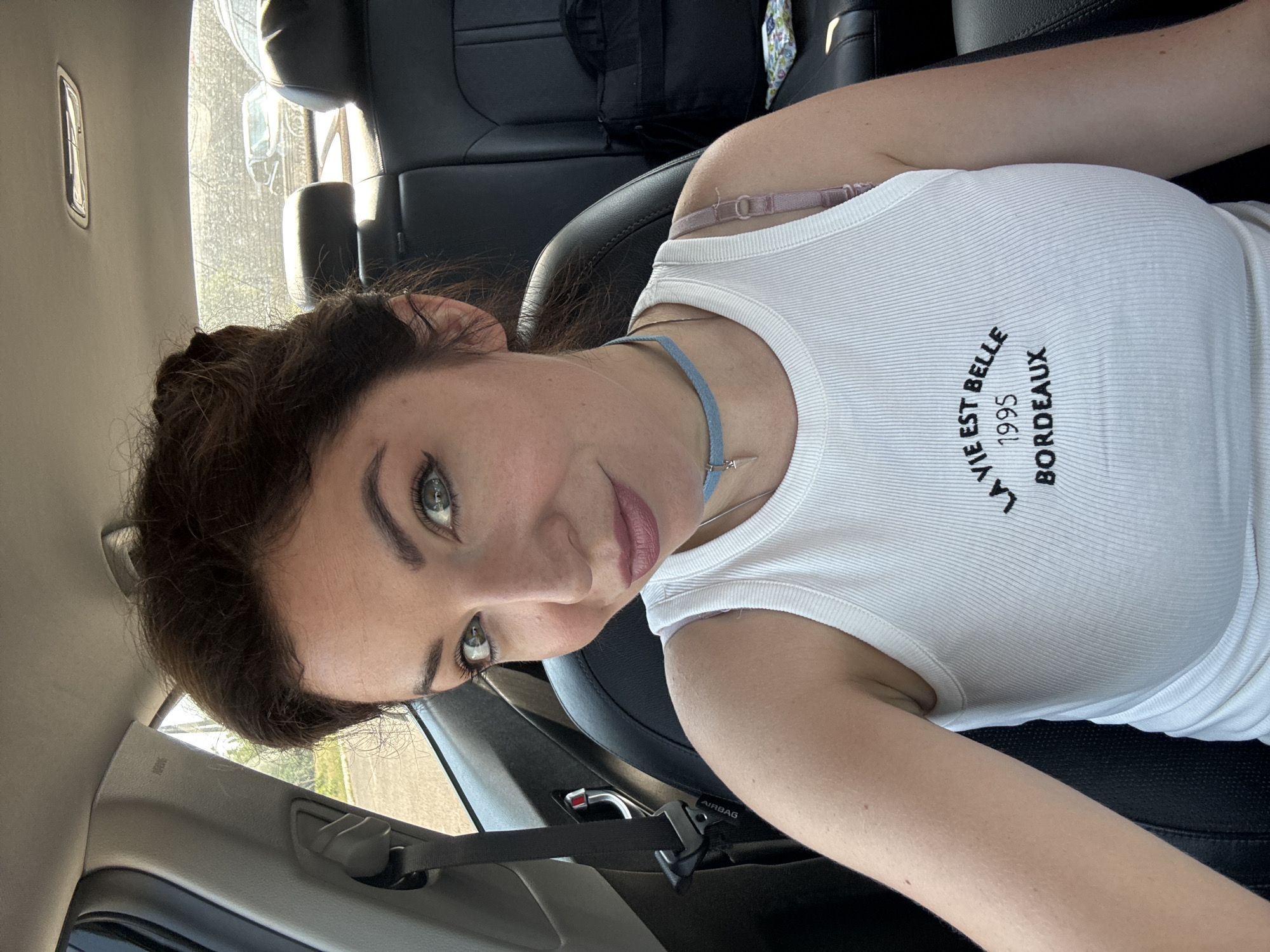 A woman with dark hair and green eyes is sitting in the passenger seat of a car, looking at the camera. She is wearing a white ribbed tank top with black text that reads "LA VIE EST BELLE 1995 BORDEAUX", a light blue choker necklace with a silver charm, and a pink bra strap is visible. The interior of the car is visible, including black leather seats and a seatbelt. The background outside the car appears to be a sunny day with trees and other cars. The exact location in Kryvyi Rih, Ukraine cannot be determined from the image.