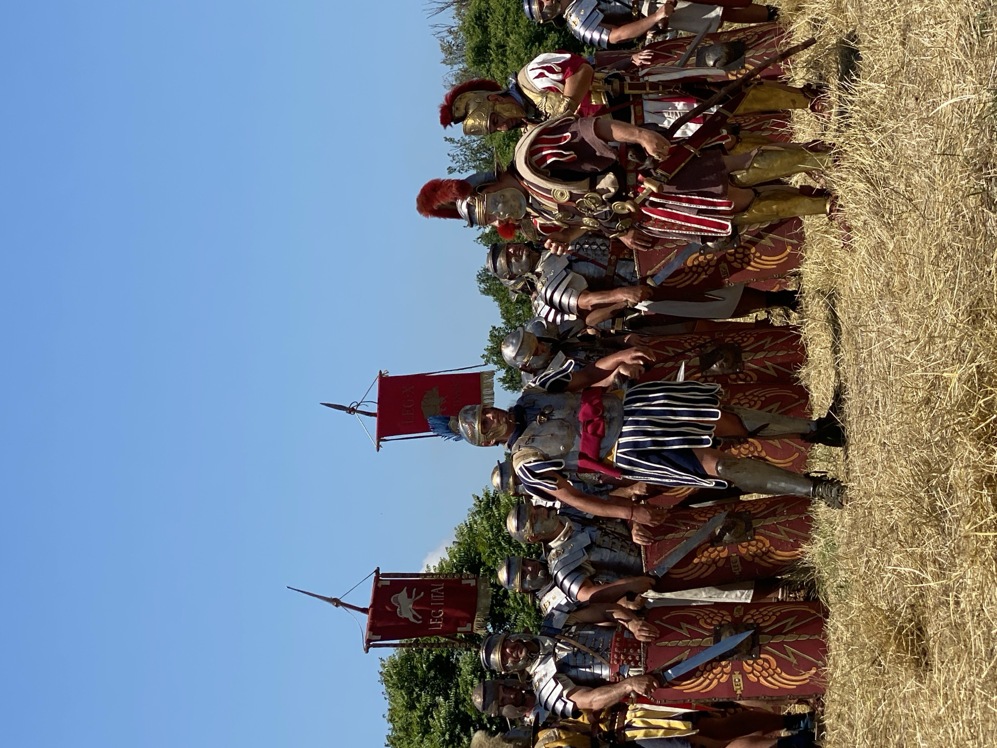 The image captures an outdoor historical reenactment in Gigen, Bulgaria, featuring approximately 15-20 individuals costumed as Roman soldiers, standing on dry, golden-brown grass under a clear, bright blue sky.
In the foreground and midground, a group of men is depicted. Most are clad in polished silver-grey segmented plate armor (lorica segmentata), various styles of helmets (galea), some with faceguards, and brightly colored tunics and pteruges (skirt-like strips). Many hold or have sheathed short swords (gladius) and are positioned behind large, rectangular, curved shields (scutum) that are predominantly red with golden winged lightning bolt or eagle-like designs.
Two central figures stand out. On the center-right, a man in a dark red tunic with white trim, red and white striped pteruges, and gold-colored greaves wears an ornate gold-colored helmet topped with a prominent red transverse plume. He appears to be addressing or interacting with the figure to his left. To his left, another man in silver-grey segmented armor, dark blue and white striped pteruges, and silver-colored greaves, wears a helmet with a partial faceguard.
In the background, a line of dense green deciduous trees and bushes forms the horizon. Above the soldiers, two vertical vexilla (standards) on poles are visible. The left vexillum is red with a white/silver stylized animal (possibly a boar or wolf) and the text "LEG ITAL". The right vexillum is also red, featuring a stylized green tree/oak leaf design and the text "LEG X". Both standards are topped with spearhead finials. The scene suggests a static formation or a pause during a re-enactment event.