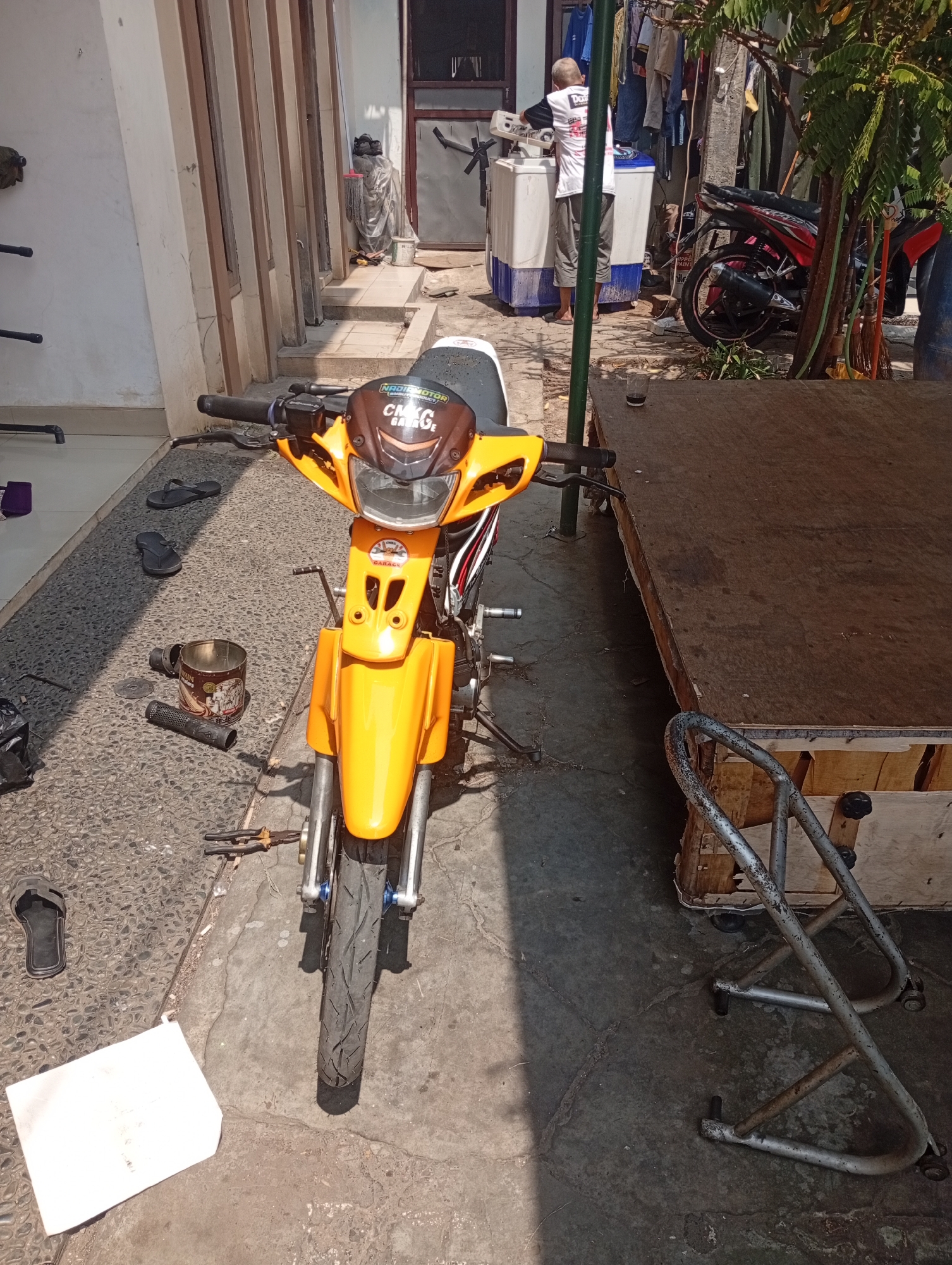 The image depicts an outdoor scene, likely a residential area or workshop, in Bekasi, Indonesia. In the foreground, a bright orange motorcycle is parked, facing forward. The motorcycle has a white seat and appears to be undergoing some maintenance or has been recently worked on, as evidenced by tools and a can lying on the ground nearby. The ground is paved with small, light-colored stones, and some cracks are visible in the concrete.
To the left of the motorcycle, a few objects are scattered on the ground, including a pair of black flip-flops, a metal can with a label, and what appears to be a part of the motorcycle's exhaust system. Further back, to the left, a section of a house with vertical wooden slats and a cream-colored wall is visible.
To the right of the motorcycle, a raised wooden platform or table is present, with a metal stand resting in front of it. Behind the platform, laundry is hanging on a clothesline, and a person in a white shirt and dark pants is standing near a washing machine, appearing to be operating it. Another red and black motorcycle is parked further back, near a tree with green foliage. The overall impression is a functional, outdoor space where activities like motorcycle repair and laundry are taking place.