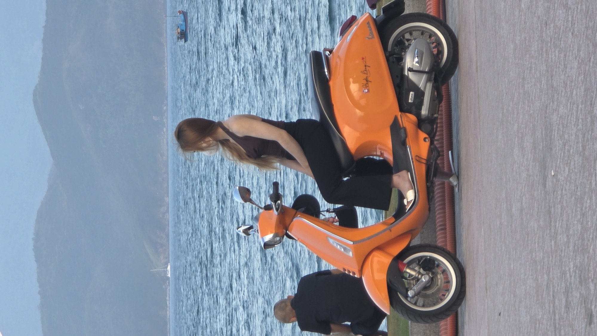 A vertical shot shows a woman leaning on an orange Vespa scooter, parked on a gray concrete slab. She is wearing a dark brown tank top and dark pants. A man is bending over the scooter near the front. In the background, a body of water extends toward mountains in the distance under a clear sky. This is likely taken in the city of Nha Trang, Vietnam given the backdrop of the ocean and mountains. A boat is seen at the back left of the frame.