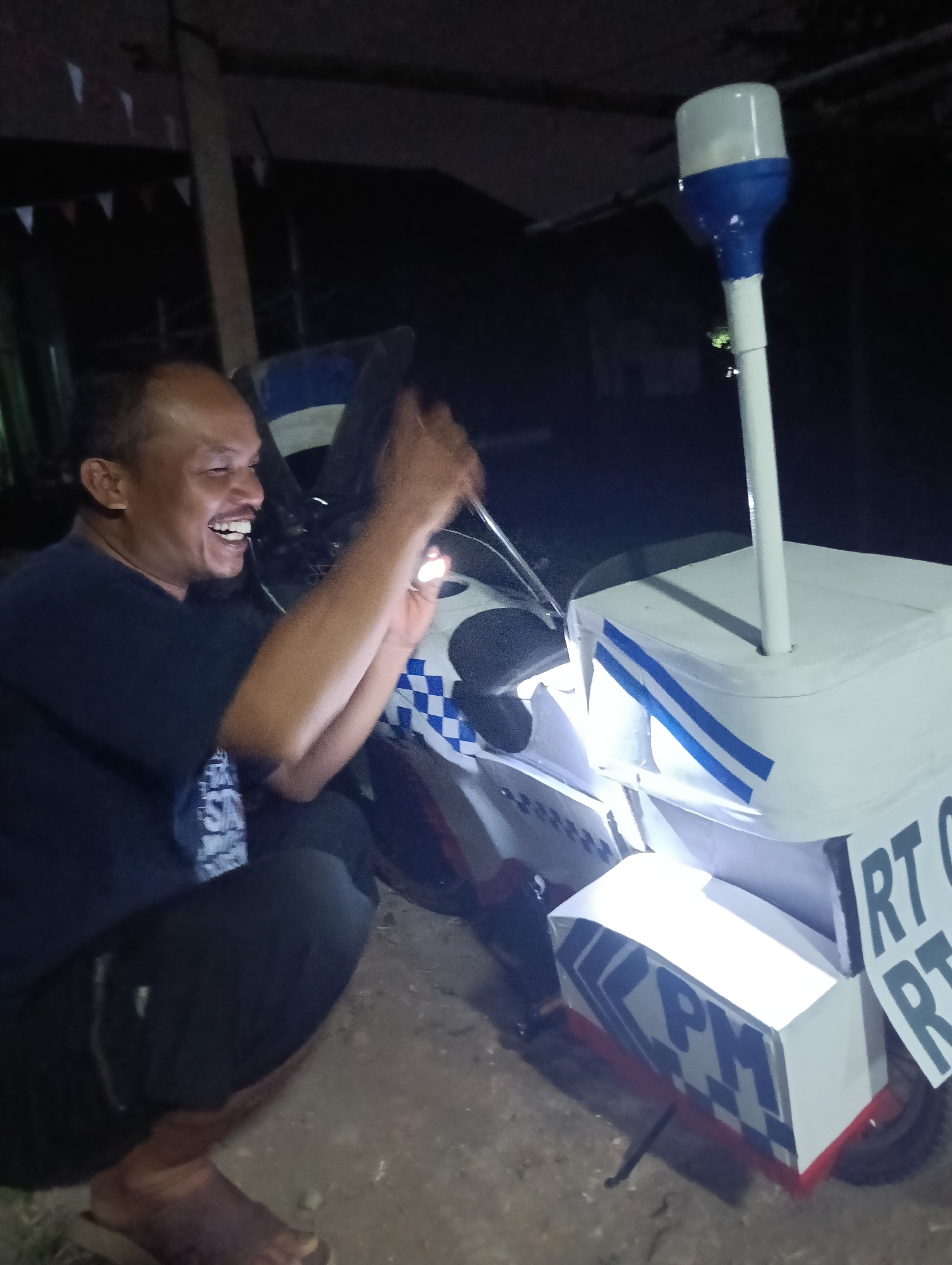 A middle-aged Indonesian man with a shaved head and a dark shirt is captured in a wide smile while squatting beside a modified motorcycle. The motorcycle has a white box-like structure attached to its rear, adorned with blue and white checkered patterns and blue stripes. A white pole with a blue and white light fixture extends upwards from the box. The man is holding a lit object, possibly a phone, and appears to be in the process of illuminating the motorcycle's structure. The scene takes place outdoors at night, with visible string lights in the background and a dirt or gravel ground. The man's body language and facial expression suggest he is engaged in a cheerful activity related to the motorcycle modification. The presence of "RT" and "RW" labels on signs attached to the box indicates it is likely related to local community organization or administration, often seen in Indonesian urban settings like Tasikmalaya.