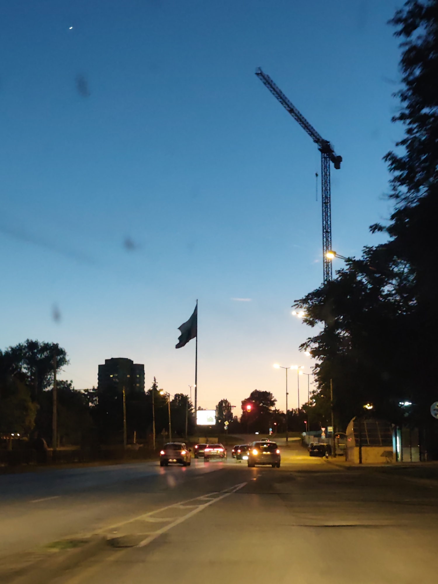 The image depicts a street scene at dusk in Sofia, Bulgaria. The sky transitions from blue to orange near the horizon. A tall crane is visible on the right side of the frame, and a Bulgarian flag flies in the distance. Cars with headlights on travel along the wet asphalt road. Streetlights illuminate the scene. Trees line the sides of the road. A building is visible in the distance behind the flag. The shot appears to be taken from inside a vehicle looking out towards the cityscape. There are some imperfections on the windshield of the vehicle. The location is likely along Tsarigradsko Shose Boulevard.