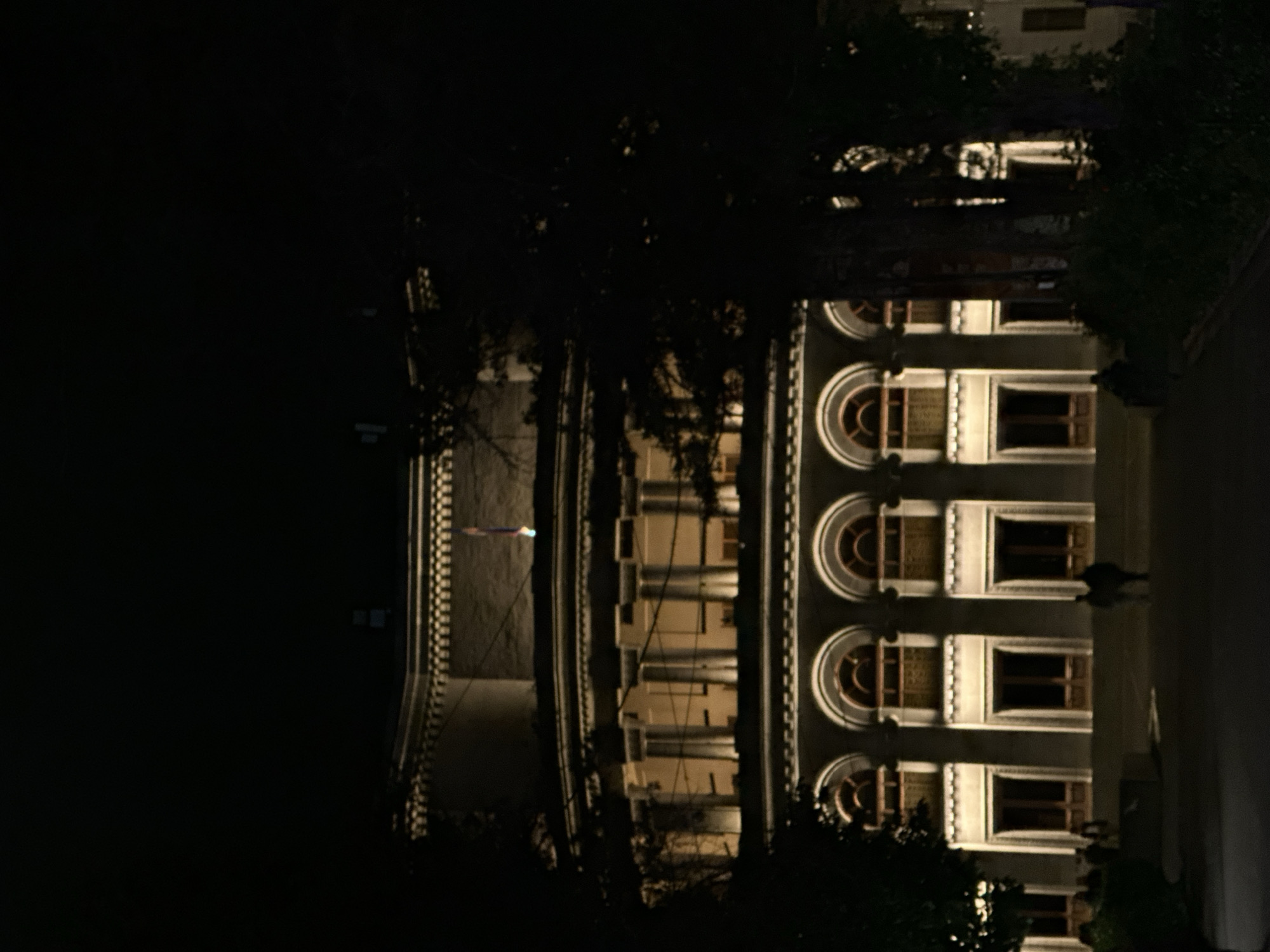 The image shows a multi-story building at night. The building has a light-colored facade with symmetrical rows of windows, some of which are arched. Lights illuminate the building's architectural details. Dark foliage is visible in the foreground, partially obscuring the view of the building. The sky is dark, suggesting it is nighttime. The image has been rotated 90 degrees counterclockwise. The location is likely Yerevan, Armenia.