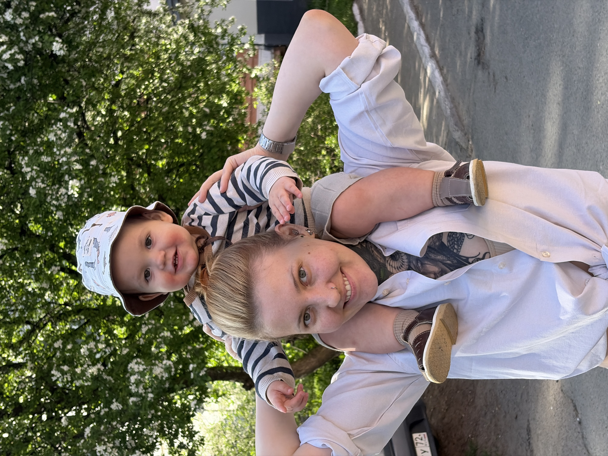 A woman is carrying a young child on her shoulders. The woman has short, blonde hair, a nose piercing, and tattoos visible on her chest. She is wearing a white button-down shirt. The child is wearing a striped long-sleeved shirt, a patterned hat, and small brown shoes. Both the woman and the child are smiling at the camera. In the background, there is a tree with white blossoms. A road and part of a vehicle license plate are visible. Based on the license plate, the image appears to have been taken in Russia, with the '72' region code possibly indicating the Tyumen Oblast area. However, given the prompt specifies the city of Chaikovsky, Russia, it's likely within that general region.