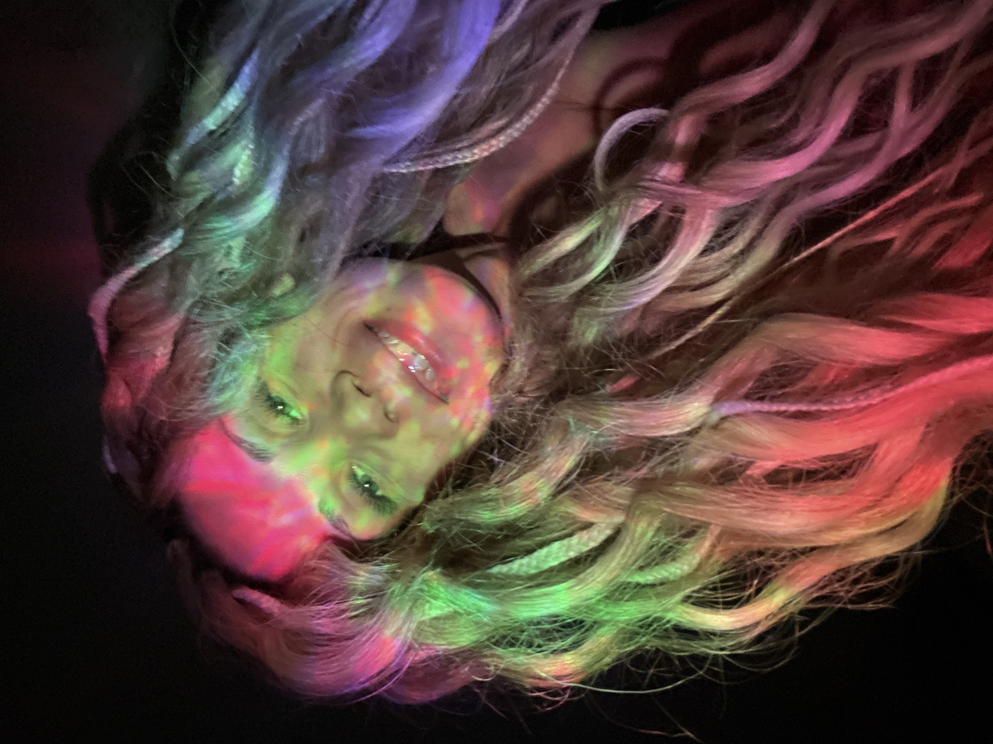 The image depicts a person with light-colored hair, illuminated by a wash of multicolored light (purple, green, red). The hair appears styled with braids. The person's face is visible, with a slight smile. The background is dark. There is no evidence to suggest this image was taken in the city of Rusanovo, Russia. The location is unspecified.