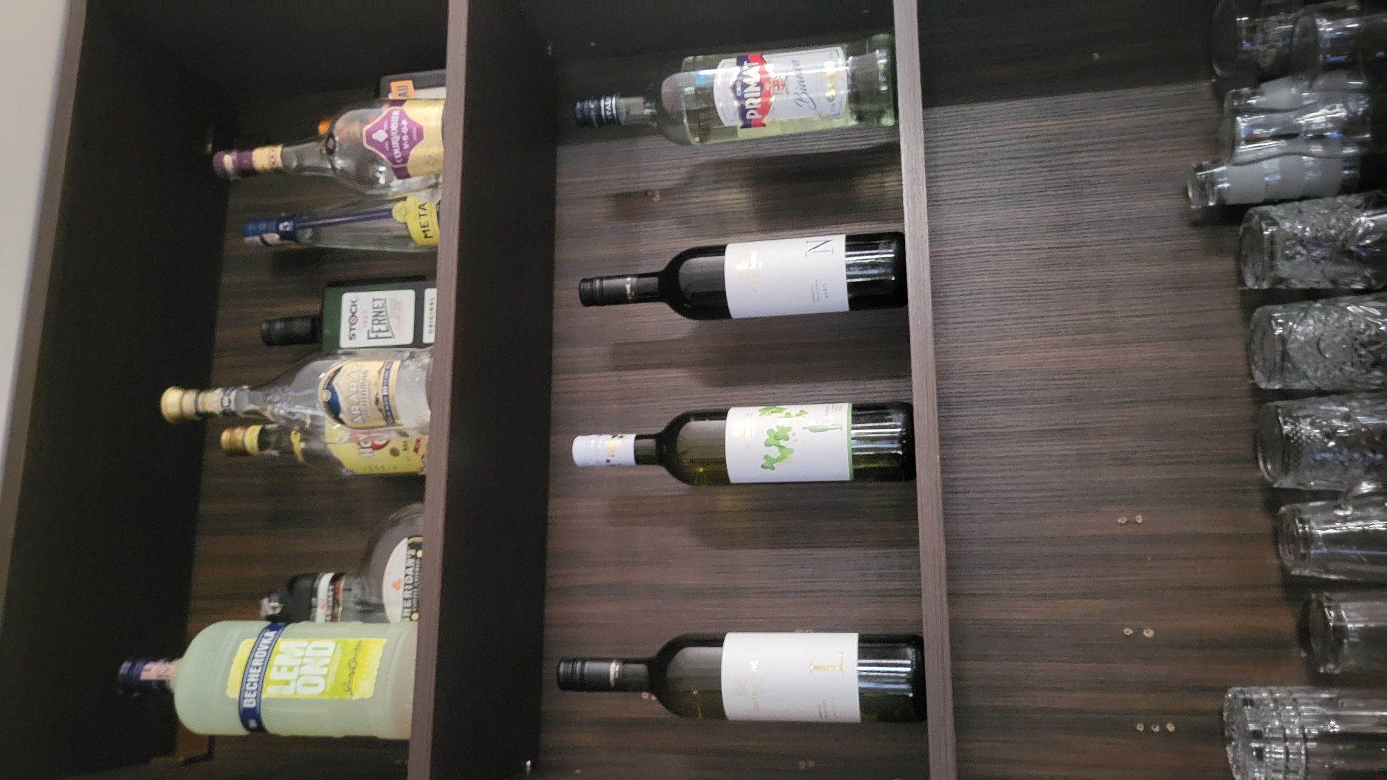 The image shows a well-stocked bar shelf in a dimly lit room, likely a home bar or a small establishment. The shelf is made of dark wood with vertical grain. On the top shelf, there are several bottles of alcohol. From left to right, a tall, light green bottle labeled "BECHEROVKA LEMOND" is prominent. Next to it is a bottle with a dark cap, followed by a clear bottle with a yellow label. Further right, there are more clear bottles, some with ornate labels, and a dark green bottle. The middle shelf displays four wine bottles. From left to right, these are a dark green bottle with a white label, another dark green bottle with a white label featuring a green leafy design, a dark green bottle with a white label, and a clear bottle with a white and red label that says "PRIMAT Bianco". The bottom shelf is mostly obscured by a row of clear drinking glasses, some with intricate patterns. The lighting suggests an indoor setting, possibly evening. The specific location in Prešov, Slovakia, cannot be determined from the image alone.