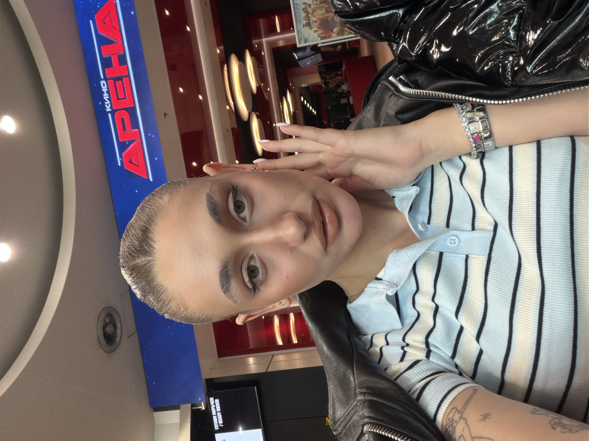 A person is taking a selfie indoors, with "Arena Kino" signage in the background. The person is wearing a striped light blue and white top, a black jacket, and a bracelet. The background shows interior architectural details, including lights and red accents. Based on the signage in Cyrillic script "КИНО АРЕНА" and the context, the location is likely within the "Arena" cinema complex. Although there are multiple Arena cinemas in Bulgaria, identifying the specific location in Sofia requires further visual context to match architectural or interior design elements visible in the image to a specific cinema location in the city.