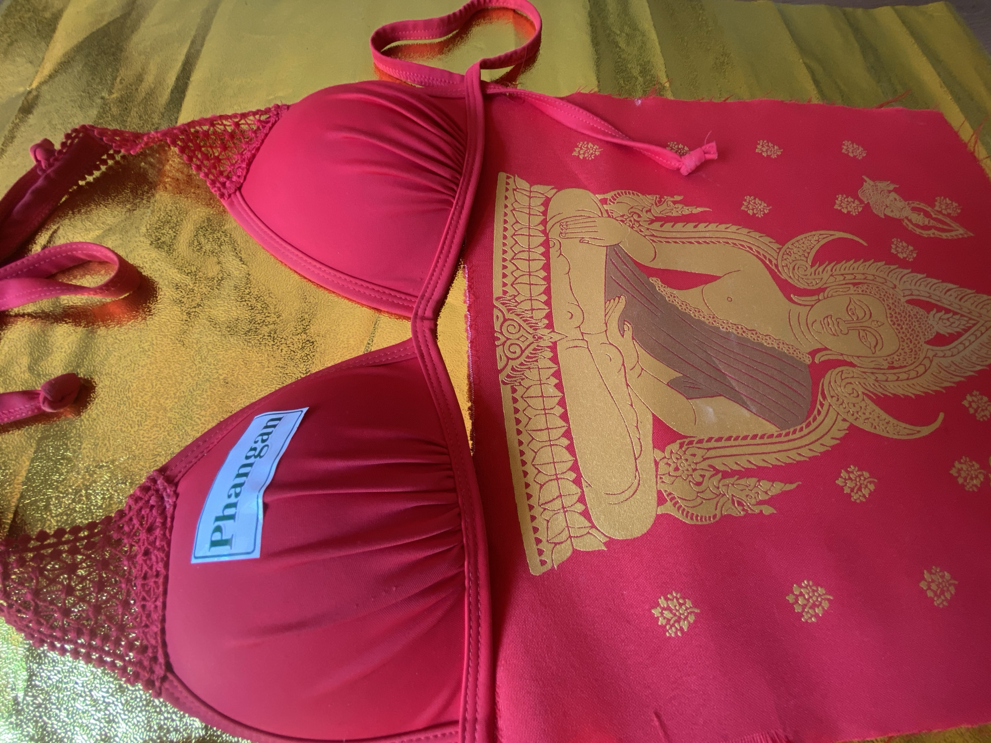 A close-up shot displays a red bikini top with lace trim, bearing a sticker with the word "Phangan" on one of the cups. It is placed on a gold-colored surface and next to a red cloth with an imprinted golden image of a figure and floral patterns. The photograph may have been taken on the island of Koh Phangan as suggested by the text on the sticker.