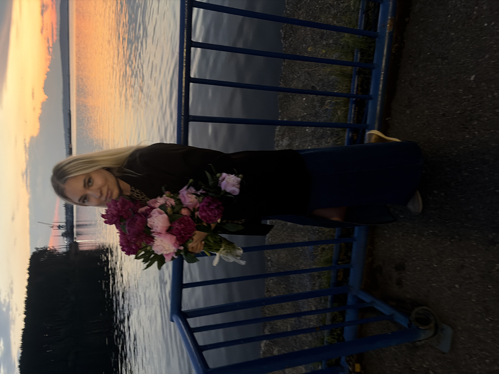 A young woman with blonde hair stands on a paved walkway next to a blue metal railing. She is holding a bouquet of pink and deep purple peonies. She is wearing a black jacket over a patterned top and a dark blue skirt. Her feet are clad in casual shoes. Behind her, a calm body of water reflects the colors of a sunset, with silhouetted trees and industrial cranes visible in the distance along the shoreline. The scene appears to be located in Chaikovsky, Russia, likely along the Kama River embankment.