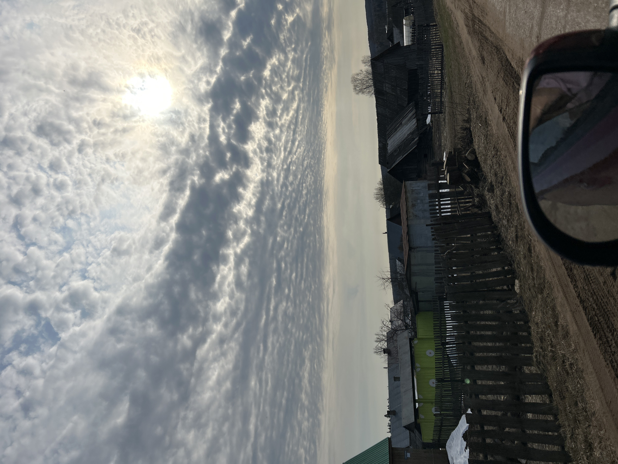 The image shows a rural scene in Уральское, Russia. The sky is filled with textured altocumulus clouds, with the sun visible through a break in the cloud cover. The landscape features a dirt road running alongside a series of dilapidated wooden structures, including houses and fences. A side-view mirror of a vehicle is visible in the foreground, suggesting the photo was taken from inside a car. Remnants of snow are visible in the lower-left corner.