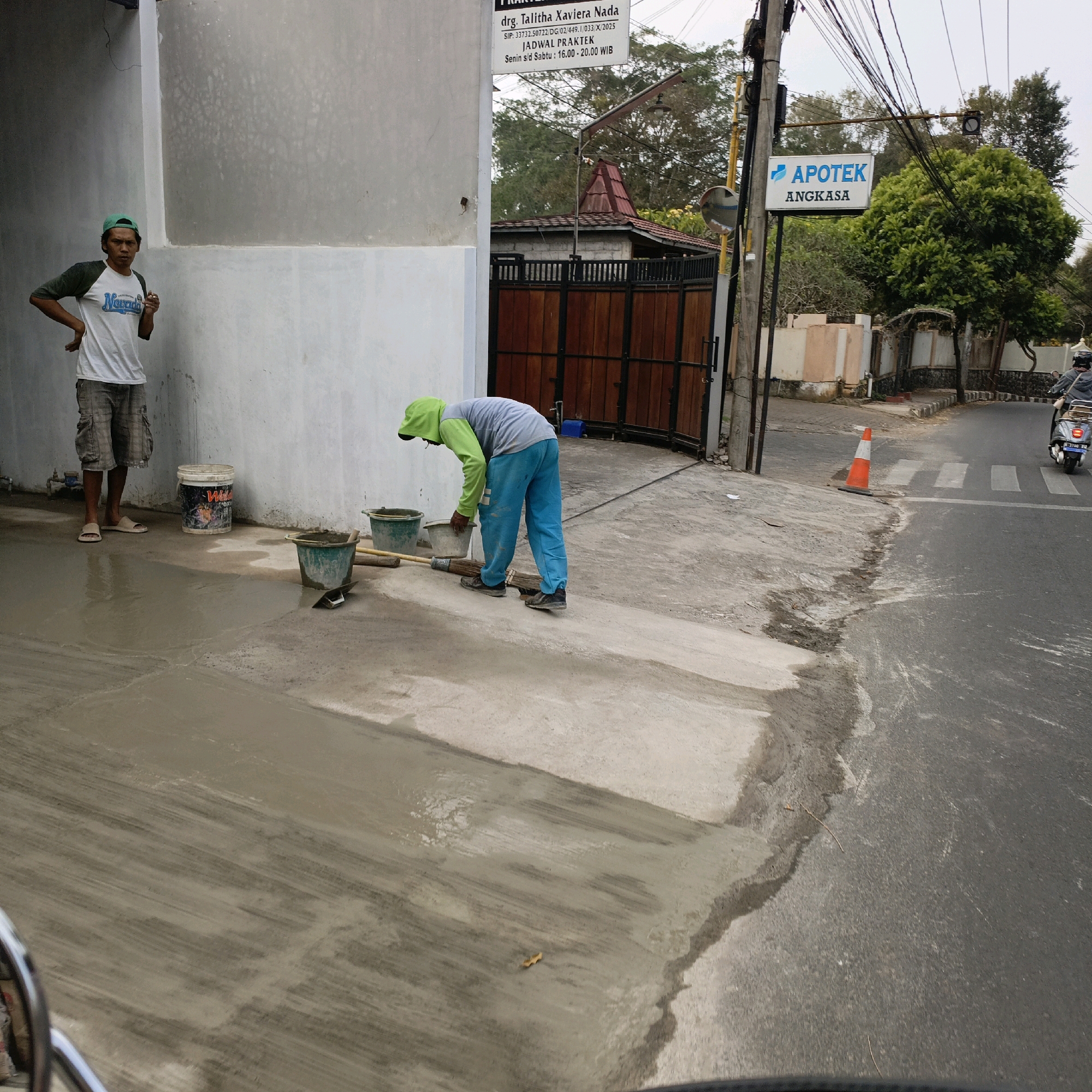 This image captures a scene in Salatiga, Indonesia, likely in front of a building that houses "APOTEK ANGKASA" and a medical practice by "drg. Talitha Xavieria Nada." The ground in the foreground is freshly laid wet cement, showing trowel marks and a slightly uneven finish. Two men are present. One man, wearing a green cap, white t-shirt with dark sleeves, and grey cargo shorts, stands with his left hand on his hip, facing towards the viewer. He appears to be supervising or taking a break.
The second man, dressed in a green hooded jacket with light blue pants, is actively engaged in working with the cement. He is bent over, holding a bucket in his left hand and appears to be using a tool, possibly a trowel or broom, with his right hand to spread or smooth the wet cement. Two green buckets and a larger white bucket labeled "Wild" are positioned nearby. A wooden broom and a small trowel are also visible on the cement.
In the background, a wooden gate with dark vertical planks and a smaller brown gate are visible, leading to a building. Beyond that, trees and overhead power lines are present. A traffic light and a sign for "APOTEK ANGKASA" are mounted on a pole on the right side of the image. A traffic cone, orange and white, is placed on the edge of the road. A motorcycle with a rider is seen in the background on the asphalt road, which features a pedestrian crossing. The overall impression is a construction or maintenance activity taking place on a street in Salatiga.