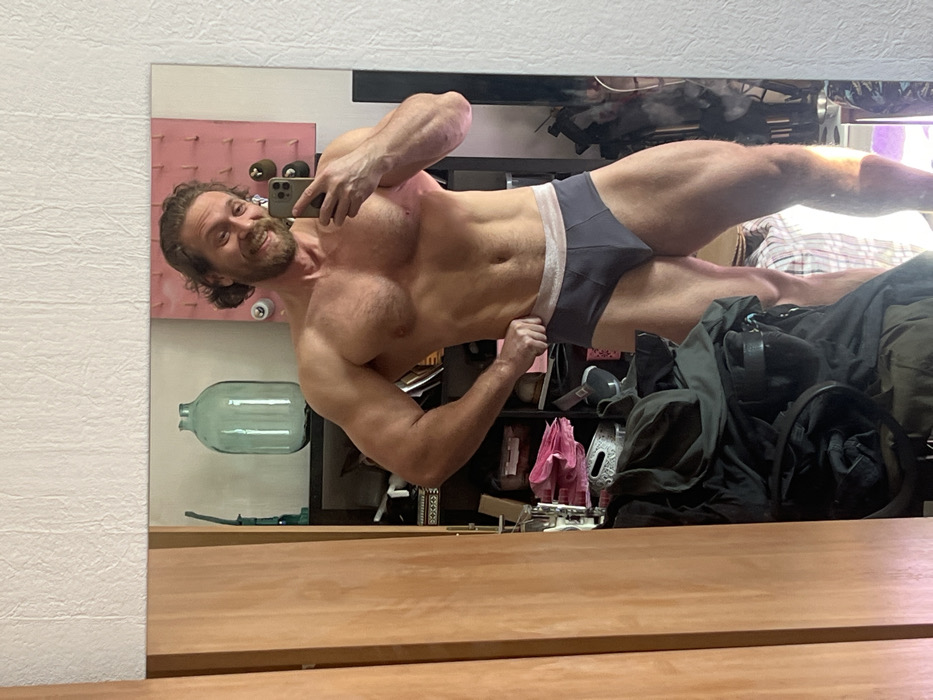 A male individual is depicted in a mirror selfie within an indoor setting in Saint Petersburg, Russia. The man, appearing to be of muscular build, stands shirtless, wearing dark grey briefs with a light-colored waistband. He possesses brown, wavy hair and a beard, and exhibits a smiling facial expression. His right hand holds a dark-colored smartphone, positioned to capture the reflection, while his left hand rests on his left hip. The background, visible in the mirror's reflection, includes a light-colored, textured wall above the mirror, a wooden cabinet/door frame to the left, and a dark shelving unit behind the individual. On the shelves, a large, translucent light green glass carboy is prominent, alongside a pink panel with several small, rounded attachments, and various miscellaneous items including what appears to be folded clothing or fabric, a white woven basket, and other dark objects. A pile of dark clothing or fabric is reflected in the lower center of the mirror, partially obscuring the lower shelves. Natural light illuminates the scene, casting shadows.