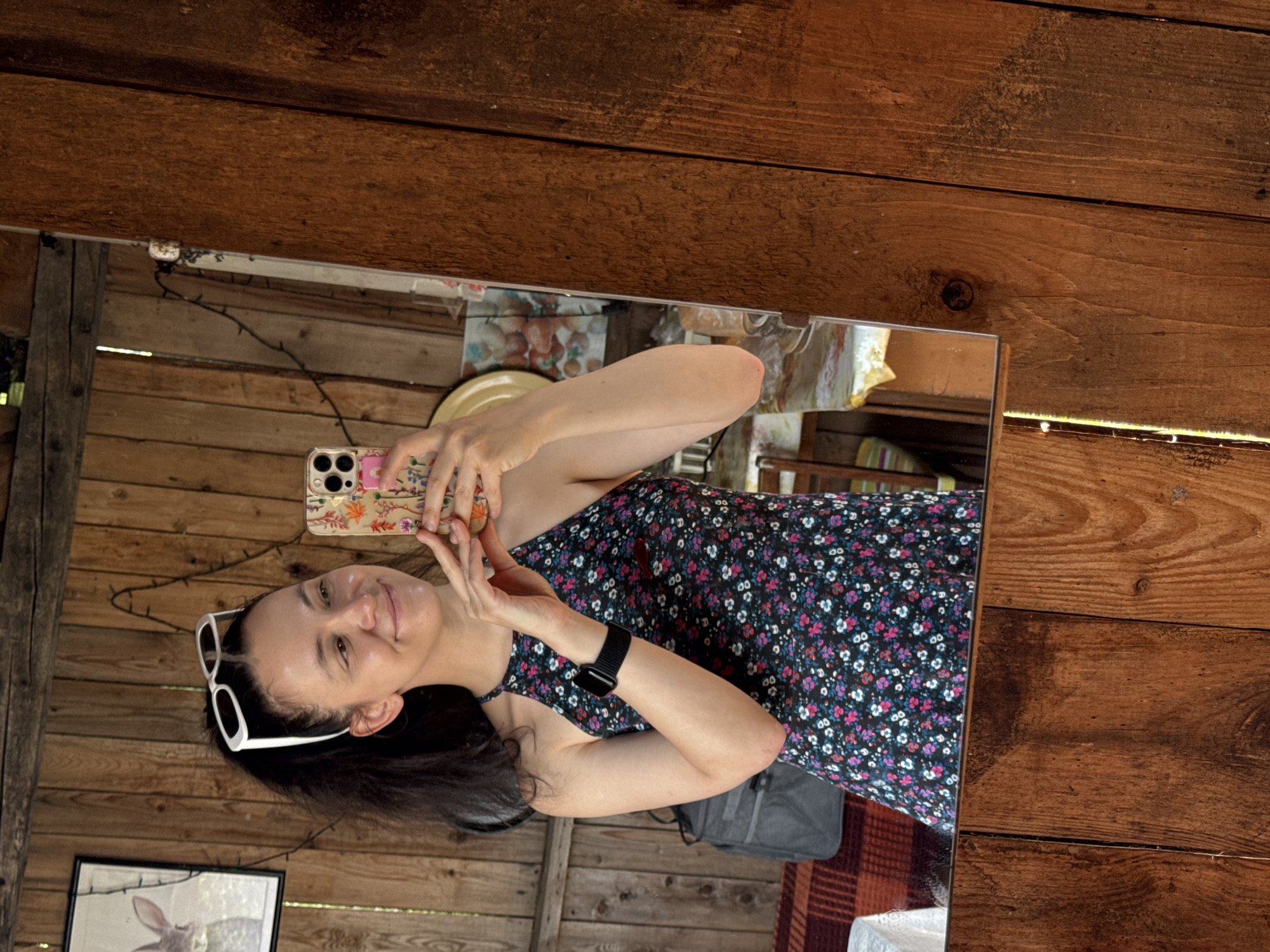 A woman with dark hair pulled back, wearing white-framed sunglasses pushed up on her head, takes a mirror selfie. She is dressed in a dark, floral-patterned halter dress with pink and white flowers. A black smartwatch is visible on her left wrist. She is standing in what appears to be an outdoor shed or rustic room with unfinished wooden walls. In the reflection, a framed picture of a rabbit can be seen on the wall behind her, as well as a string of fairy lights. There is a grey bag on a table covered with a red and white checkered cloth. The location is not identifiable as Zasechnyi, Russia.