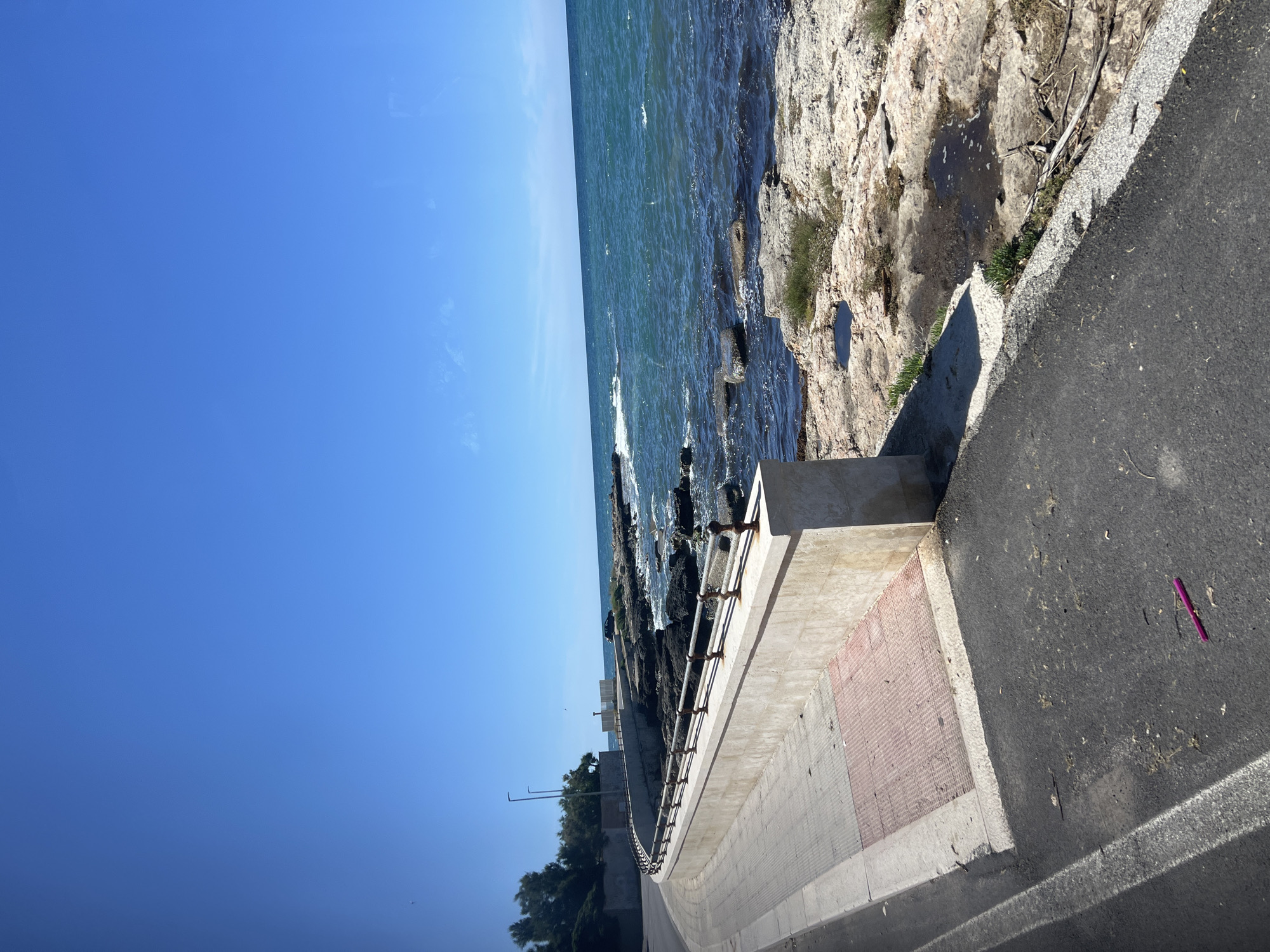 The image captures a sunlit coastal scene, likely a promenade or road alongside the sea in Bari, Italy. The upper portion of the frame is dominated by a clear, bright blue sky with only minimal, faint white clouds, indicating excellent weather.
To the right, the deep blue-green waters of the sea stretch out, marked by gentle waves and some white foam breaking against a rugged, light-colored rocky shoreline. The rocks are irregular, and small patches of green vegetation cling to them.
A concrete retaining wall or barrier separates the rocky coast from a paved area on the left. This wall features a metal railing, painted a reddish-brown, which follows the curve of the path into the distance. Part of the concrete wall also has a textured, reddish-brick-like pattern.
In the foreground and extending into the middle distance on the left, a dark asphalt road or promenade is visible, bordered by a lighter concrete curb. Distinct shadows fall across the asphalt and concrete structures, suggesting bright sunshine, likely during the late morning or afternoon. On the asphalt in the foreground, a small, bright pink or purple object lies on the ground. Further down the curving path, several street light poles and distant green trees can be seen.
The scene is tranquil and devoid of people or specific activities. There is no visible text in the image.