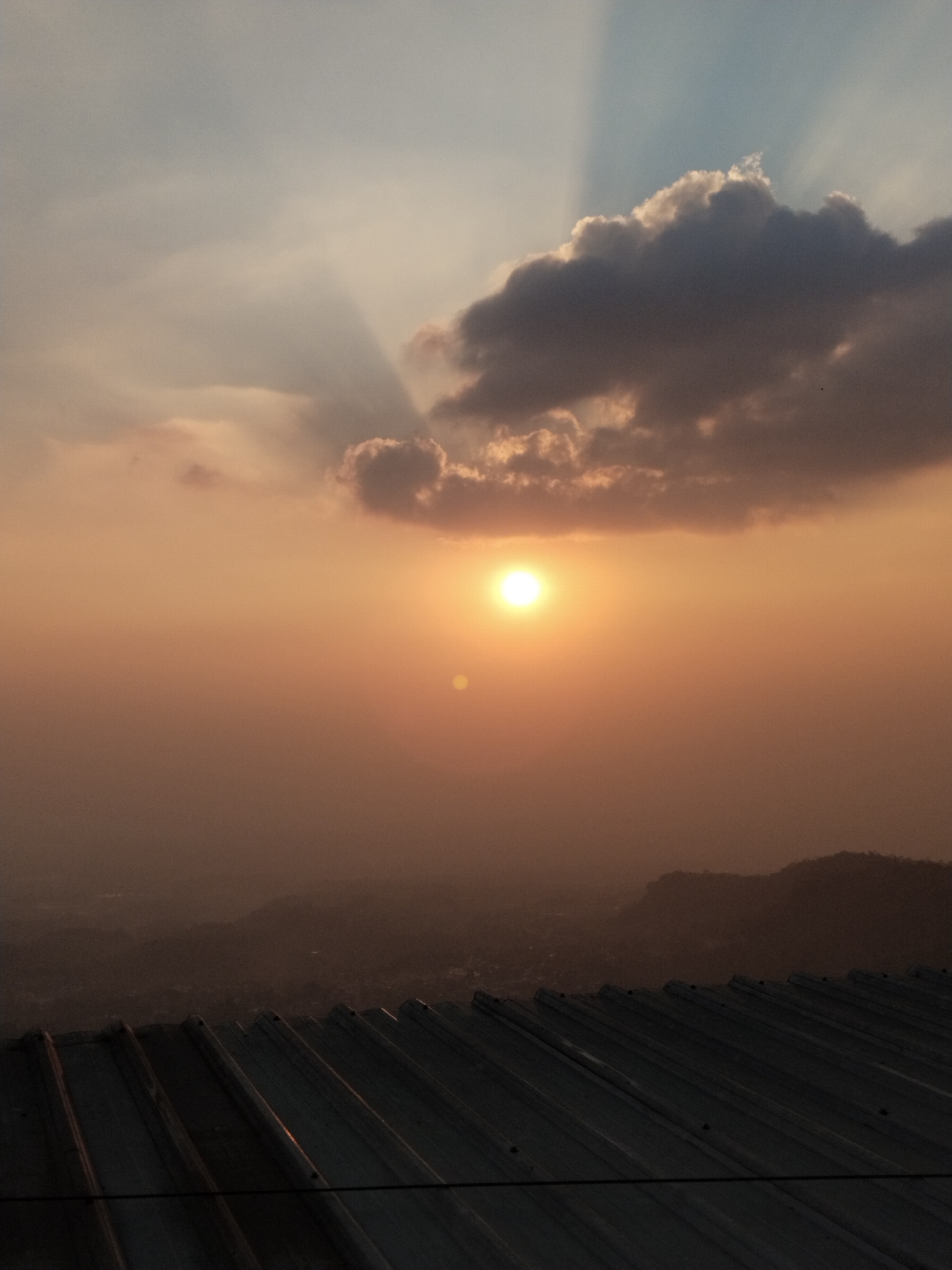 The image depicts a sunset view from a rooftop in Gumeng, Indonesia. The sky is dominated by warm orange and yellow hues, with the sun positioned in the upper-center, casting a bright glow. Rays of light emanate from behind a dark cloud formation in the upper portion of the sky. In the foreground, a corrugated metal roof is visible, angled upwards from the bottom of the frame. Beyond the roof, a hazy landscape of hills and distant city lights can be seen, suggesting an elevated viewpoint overlooking the town of Gumeng. The overall atmosphere is serene, with the fading sunlight creating a dramatic and atmospheric scene. There are no people or animals visible in the image.