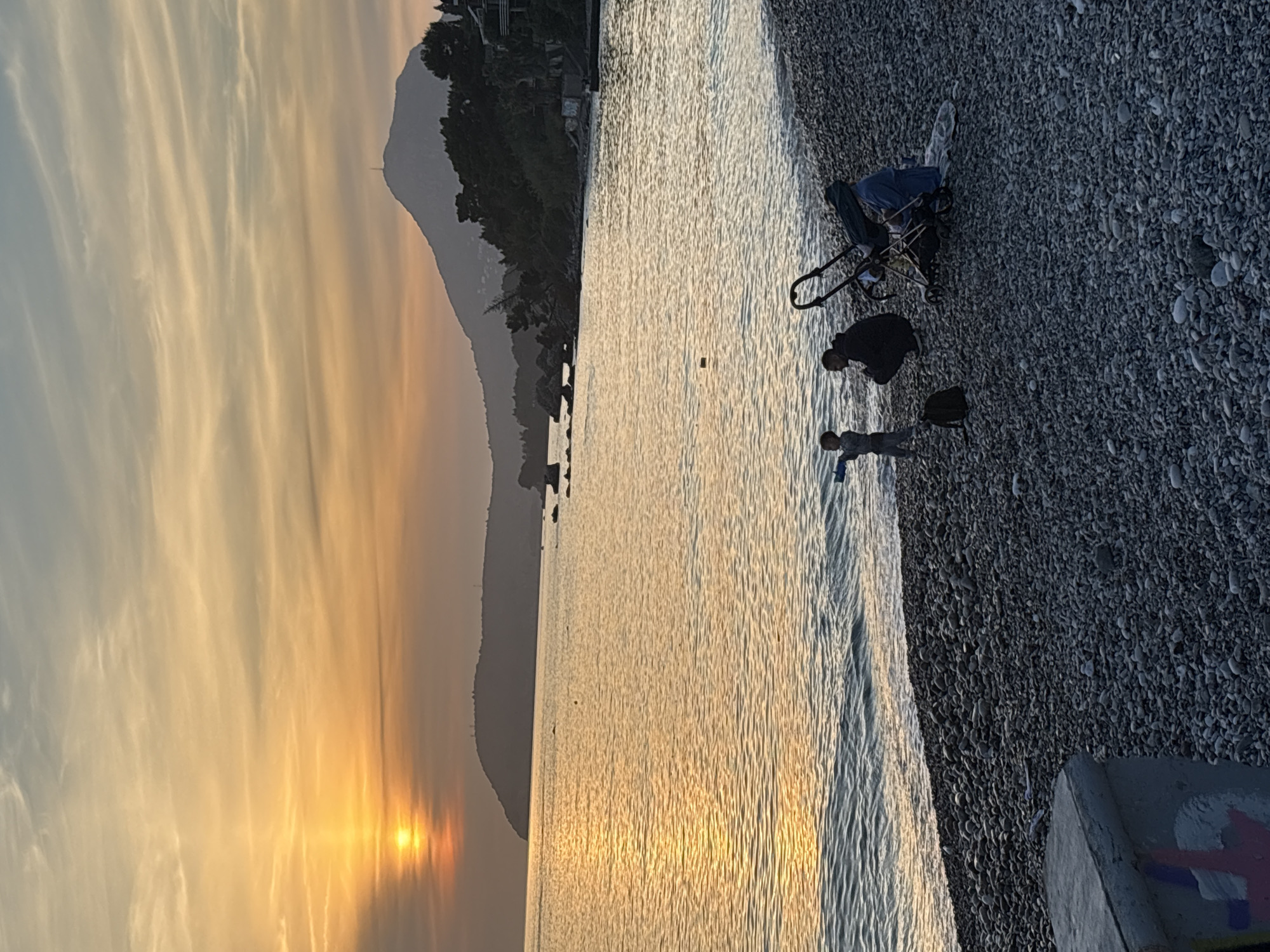 The image depicts a beach in Bar, Montenegro at sunset. The sea reflects the golden light of the setting sun. A mountain range is visible in the background. On the pebble beach, a child is near the water, with an adult standing behind them. A stroller and a bag are on the pebbles. A concrete block with graffiti is in the foreground.