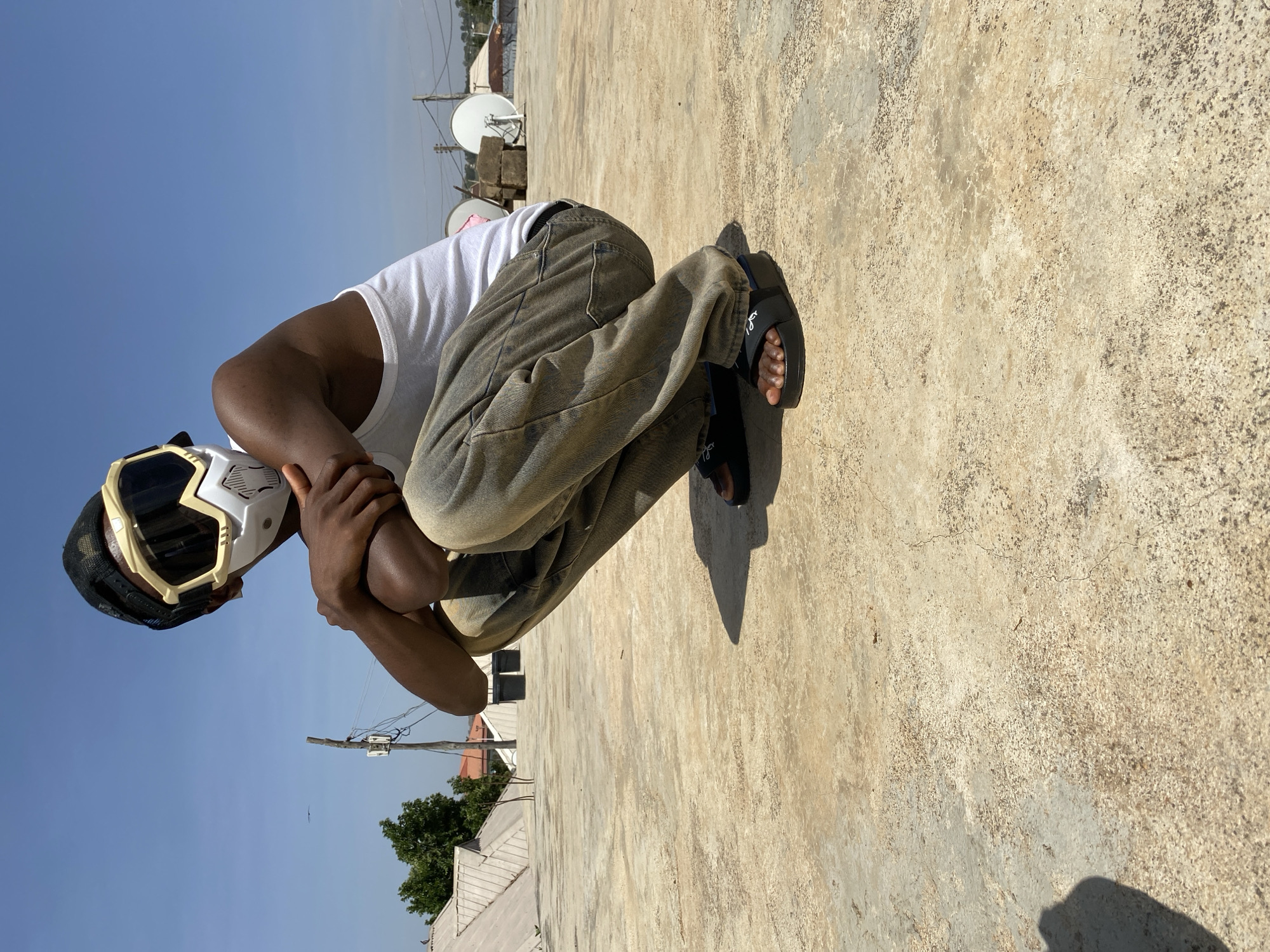 A person is crouched on a flat, concrete rooftop under a clear, blue sky in Bukuru, Nigeria. The individual wears a white tank top, mottled brown pants, and black sandals. They have a black baseball cap and are wearing a white and gold-trimmed face mask. Their arms are crossed, and they are looking downward. In the background are various buildings, satellite dishes, and utility poles typical of an urban environment. The rooftop is cracked and weathered, and the sunlight casts a shadow of the person onto the surface. The lighting suggests it is daytime, with the sun positioned behind the person.
