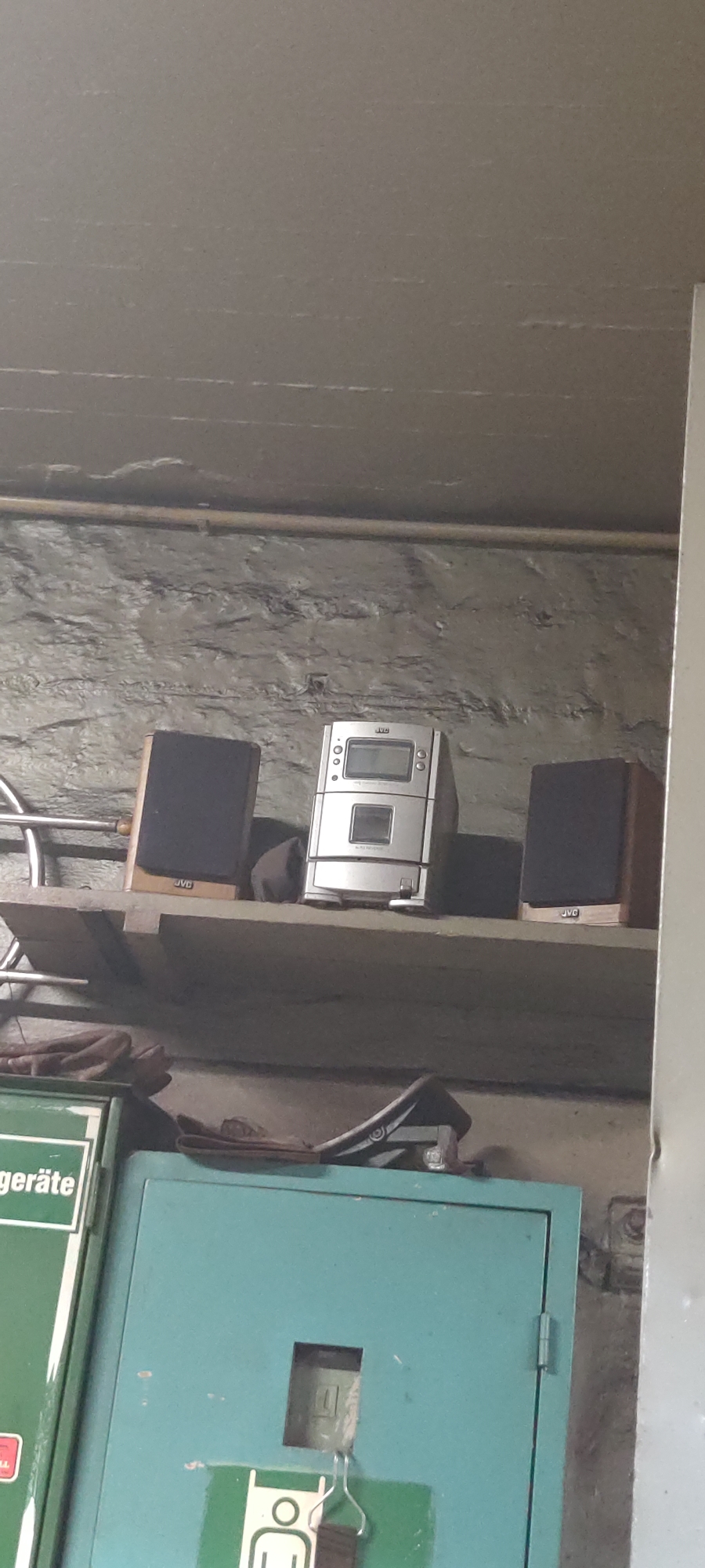 This image depicts a storage area with a concrete ceiling and stone walls. A wooden shelf is mounted on the wall, holding several electronic devices. On the left side of the shelf, there is a black speaker with a wooden front featuring the "JVC" logo. To its right is a silver JVC stereo system with a digital display and a CD player. To the right of the stereo is another black speaker with a wooden front, also bearing the "JVC" logo. Below the shelf, there is a green metal cabinet with a white label that reads "gerate" (which is likely part of a larger German word like "Messgeräte" or "Gerätehaus"). The cabinet has a hinged door with a square cutout and a metal hook. The overall scene suggests a utilitarian space, possibly a workshop, garage, or storage room. The specific location in Salzgitter, Germany, cannot be determined from the visual information alone.