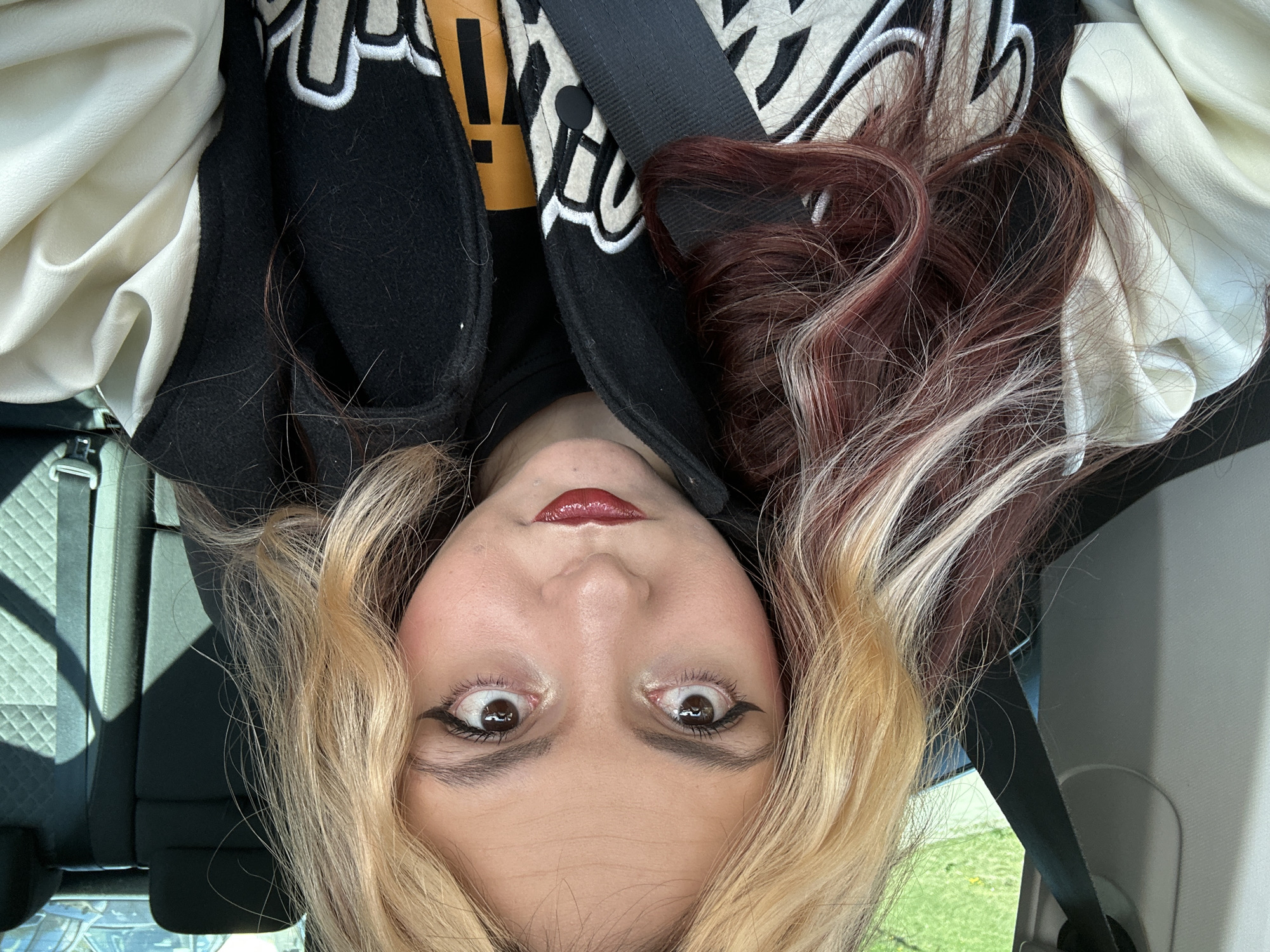A person is in a car, oriented upside-down in the frame. The person has light skin, brown eyes with black eyeliner, and red lipstick. Their hair is multicolored: blonde, burgundy, and white-blonde. They are wearing a black letterman-style jacket with a white leather sleeve and a yellow and white design on the body. A black seatbelt is visible across the person's chest. The background shows a car seat and a glimpse of green foliage outside the window. The city of Sofia, Bulgaria is not discernible from the image.