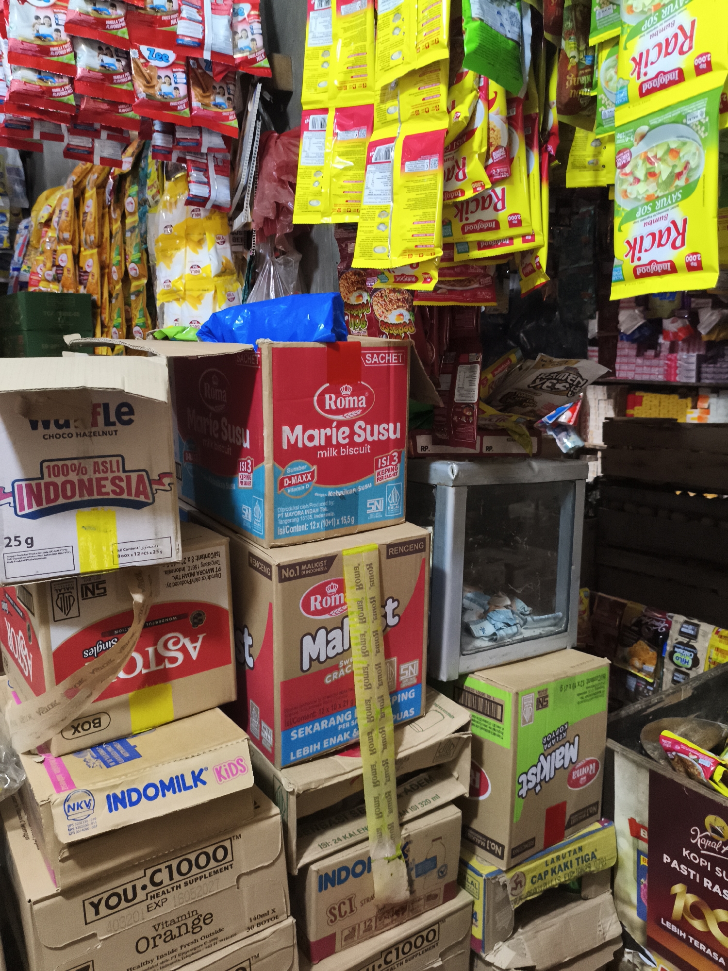 The image shows a cluttered display of various food and beverage products inside a small shop or "warung" in Indonesia. The products include "Roma Marie Susu" biscuits, "Indomilk Kids" milk, "You-C1000" vitamin drinks, "Wafer Tango" wafers, "Racik" instant soup seasonings, and "Kapal Api" coffee. These items are stacked in boxes and hang from above, creating a visually dense scene. The location is identified by labels on the packaging, indicating manufacturers and distributors operating in the Depok area of Indonesia. There are no people visible in the frame, and the scene is still and not in motion. The environment appears to be dimly lit with poor illumination from the ceiling and items hung up.