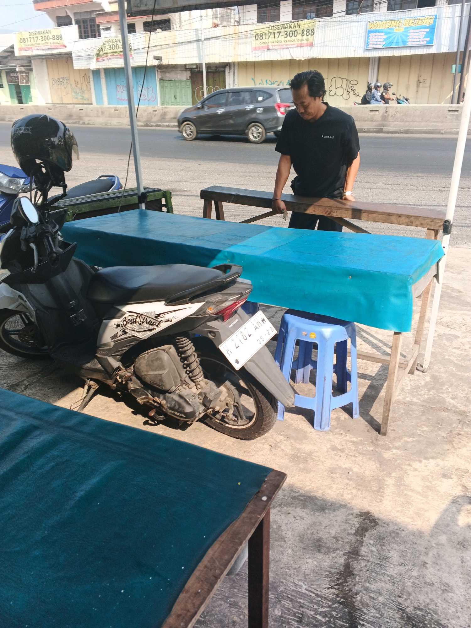A man in a black t-shirt and black pants stands behind a wooden table covered with a blue cloth, holding a set of keys in his right hand. A blue plastic stool is positioned beneath the table. To the left of the table is a motorcycle, a helmet resting on its handlebars. In the background, a dark-colored car drives on the street, and buildings with various signs line the opposite side of the road. A concrete barrier separates the road from the sidewalk area where the man and the table are situated. The scene appears to be outdoors, with sunlight casting shadows on the ground. The signage in the background, "DISEWAKAN 081717-300-888" and "WARUNG REJEKI," suggests the location is in Ungaran, Indonesia, likely a commercial or street-side area.