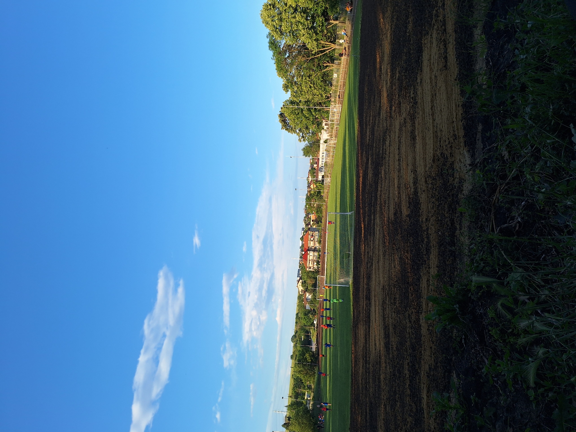The image captures a wide-angle view of a sports complex, specifically a football pitch, situated in Ezerovo, Bulgaria, under a clear blue sky with scattered white cumulus clouds. The foreground consists of a dark, unpaved, uneven earth area showing distinct horizontal striations, with sparse green vegetation emerging from the soil.
The midground features a vibrant green grass football pitch, illuminated by late afternoon or early evening sunlight, casting long shadows. On the pitch, numerous individuals, primarily males, are actively engaged in a football game or practice. They are clad in distinct sportswear: several players in blue, some in orange, others in red, and one individual identifiable as a goalkeeper in a bright green kit positioned near the goal. A white-netted football goal stands prominently on the left side of the pitch, closer to the viewer.
Beyond the pitch, in the background, a residential area is visible, comprising various buildings with tiled roofs, some orange/red and others lighter in color. A dense line of mature green trees, particularly on the right side, separates the pitch from further structures and a hilly landscape. A dirt track or path runs alongside the pitch on the right, where a few individuals are seen walking or sitting. Utility poles and overhead lines are also present, indicating infrastructure. The overall scene suggests an active community sports facility.