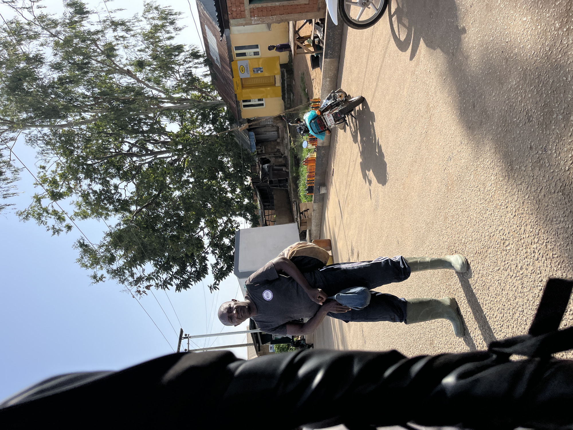 A man stands on a paved road in Bukuru, Nigeria. He is wearing a grey t-shirt, dark pants with white stripes down the sides, and green rubber boots. He has a light brown bag slung over his right shoulder and is holding a blue pouch in his left hand. The man is looking towards the camera and appears to be smiling. In the background, there is a building with a yellow facade and a corrugated metal roof, along with some trees and a motorcycle. The scene suggests it is daytime, with clear, sunny weather. The road is made of asphalt with small stones visible. The perspective is from a low angle, possibly from a vehicle. There is a logo on the man's t-shirt, but it is not clearly legible.