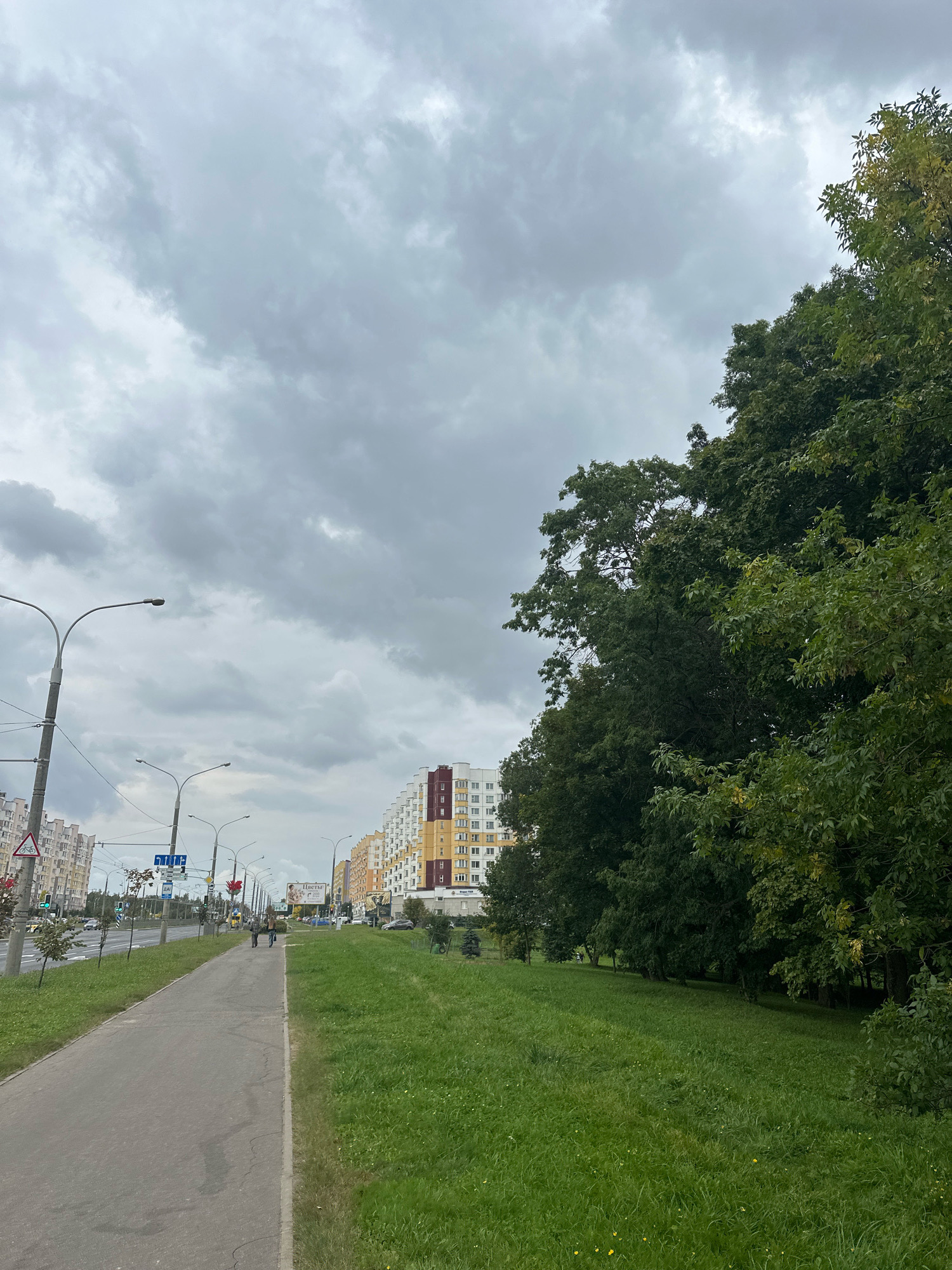 The image captures a scene on a cloudy day in the city of Minsk, Belarus. A paved walkway runs along the left side of the frame, bordered by a grassy area. Beyond the walkway, a street is visible with cars and streetlights. Multi-story residential buildings with colorful facades line the street in the background. On the right side of the frame, a dense line of green trees dominates the view, obscuring the sky. Two individuals are walking on the walkway in the mid-ground, facing away from the camera. The overall atmosphere is overcast and suggests a typical urban landscape with residential areas and greenery.