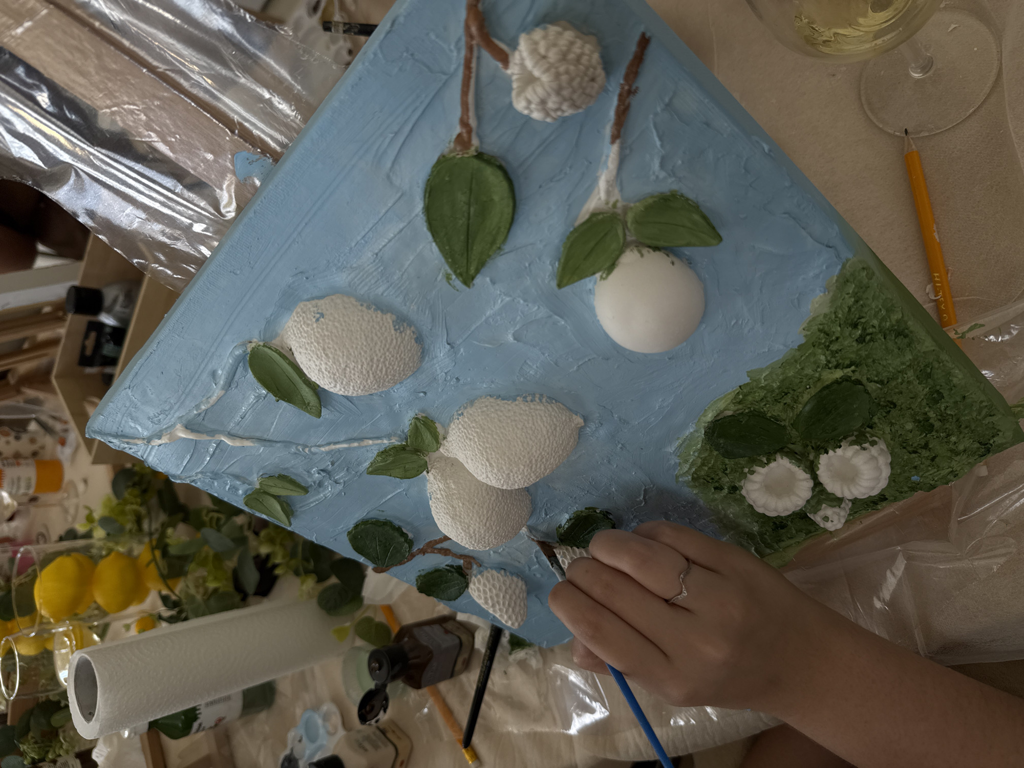 A high-angle shot shows an artist's hand holding a blue-handled paintbrush, applying paint to a triangular canvas depicting lemons on branches against a textured, light blue background. The canvas is surrounded by art supplies: a roll of paper towels, a glass of liquid (possibly white wine or water), paint bottles, and other brushes. The scene suggests an indoor setting, possibly an art studio or a home workspace. There is no evidence indicating the location in the city of Tel-Aviv, Israel.