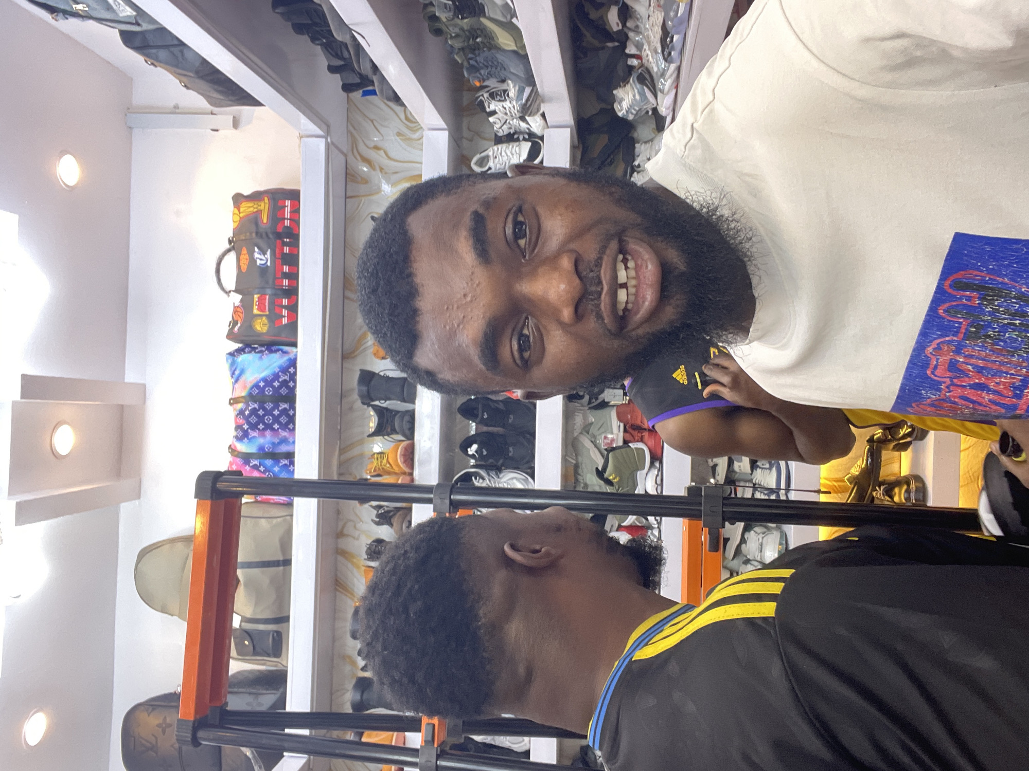 Two Black men are the primary subjects of the image. The man closer to the camera has a full beard, short hair, and is wearing a white t-shirt. He is smiling and appears to be holding up a blue sign. The second man, who is facing away from the camera, has short hair and is wearing a black shirt with yellow stripes on the shoulder. The setting is an indoor retail space with shelves stocked with handbags and shoes. The location is Jos, Nigeria.