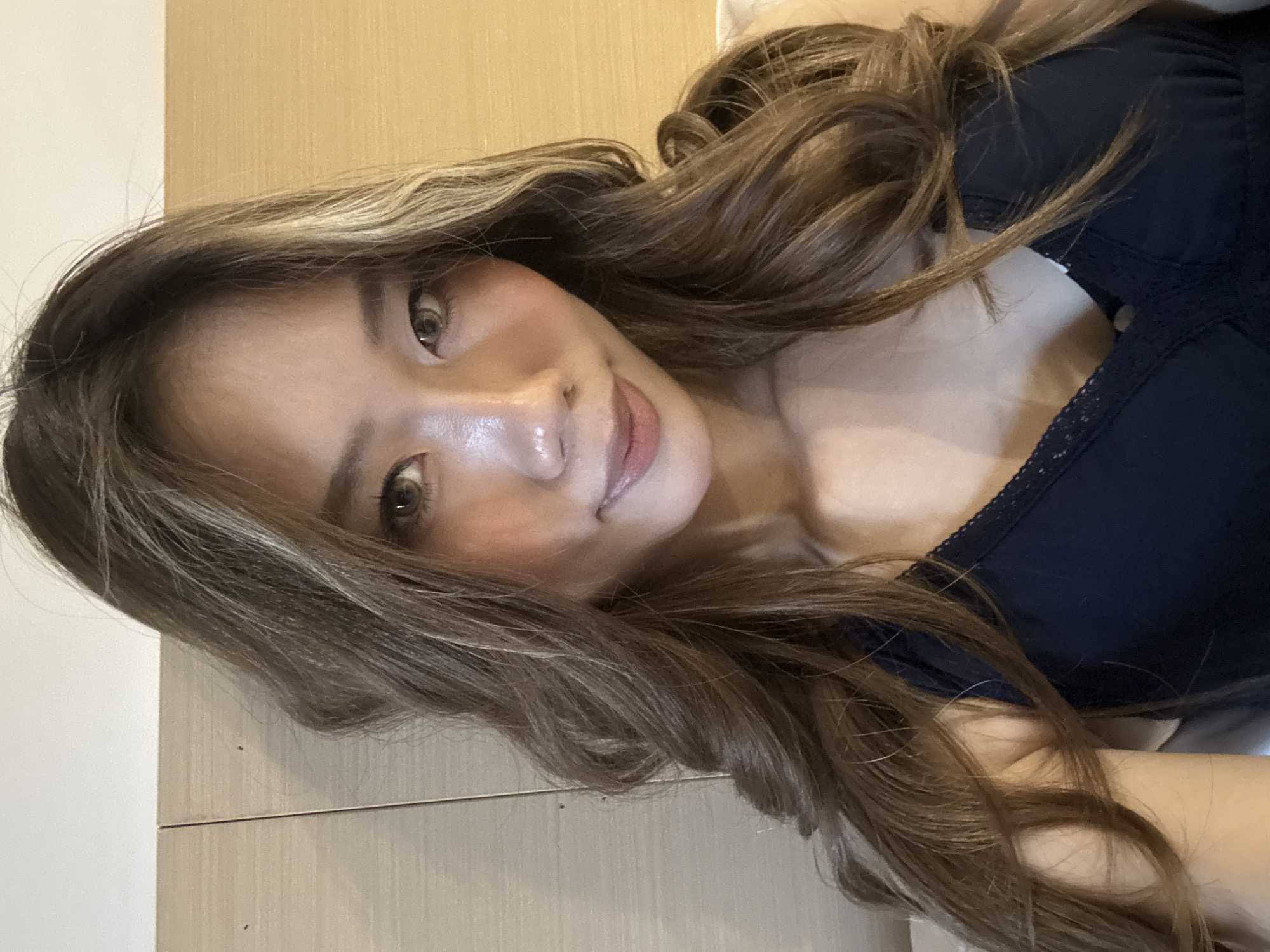 The image is a close-up portrait of an Asian woman with long, wavy brown hair that has blonde highlights. She is wearing a dark blue top with a lace-trimmed neckline. Her skin appears clear with a glossy finish. She is smiling slightly, and her eyes are open, looking directly at the camera. The background is a neutral-colored wall with a wooden paneling design. There is no evidence to determine this image was taken in Yuzhong District, China.