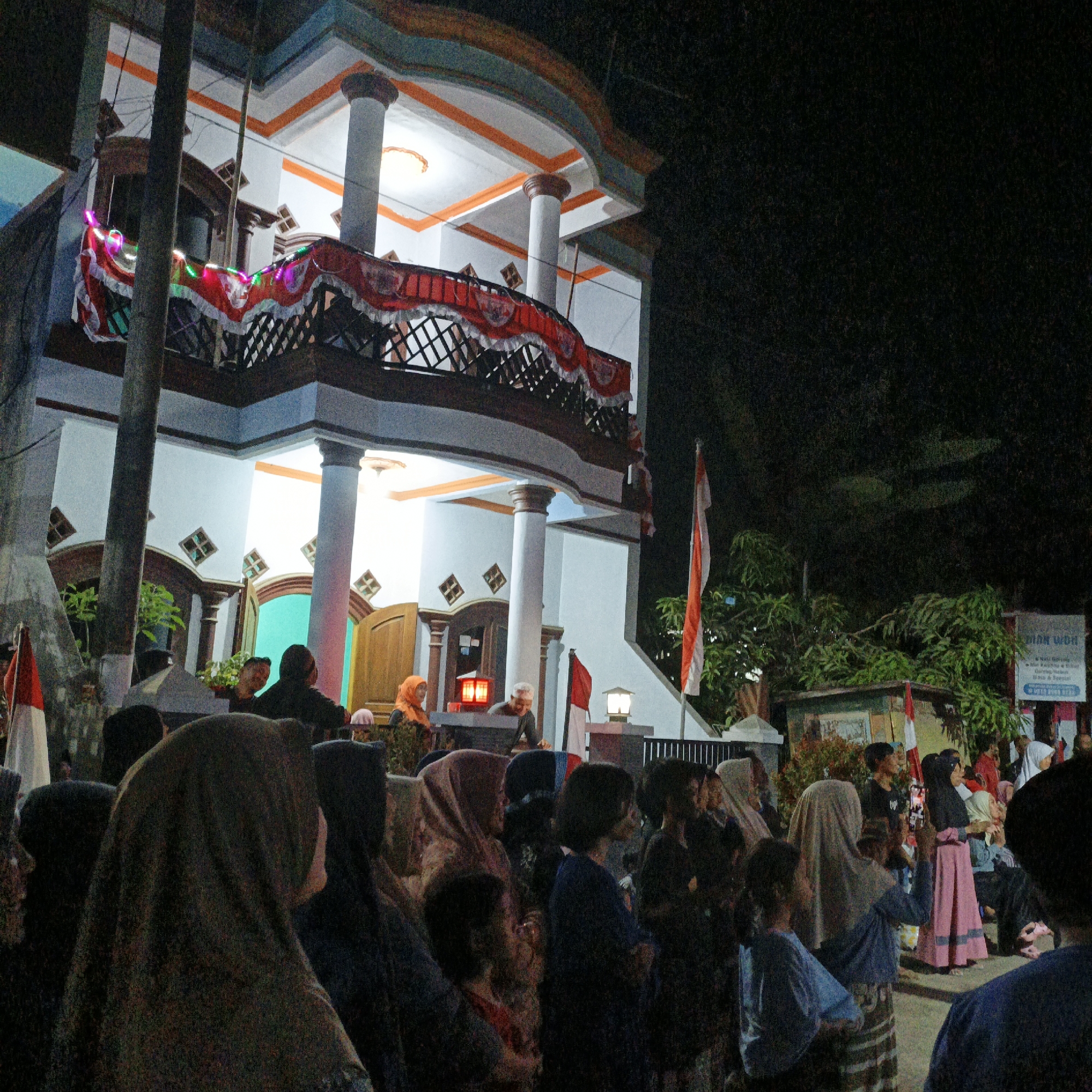 The image depicts a nighttime scene in Tanahbaya, Indonesia, likely during a festive or communal gathering. A two-story building with a balcony decorated with red and white fabric banners and string lights is a prominent feature. The building's facade is painted white with orange and blue trim. Several people are gathered in front of and around the building. In the foreground, a crowd of individuals, many wearing headscarves, are facing towards the building. Indonesian flags, red and white, are visible in multiple locations, including a large flag on a flagpole to the right of the building and smaller flags near the entrance. Several individuals are looking towards the building, with some appearing to be holding up phones or cameras, suggesting an event is taking place or being observed. The overall atmosphere appears to be one of community and celebration.