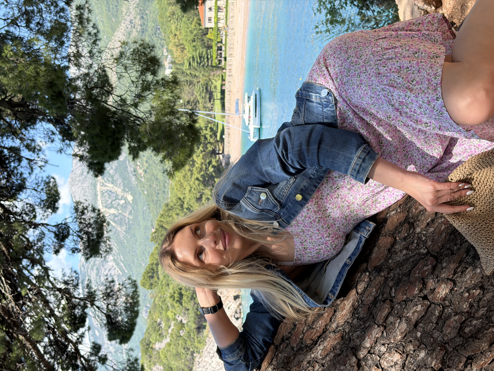 The image shows a woman with blonde hair sitting on a tree trunk in an outdoor setting, likely in or near Sveti Stefan, Montenegro. She wears a pink floral dress, a denim jacket, and a dark watch. The location overlooks a bay with turquoise water, a sandy beach, a white boat, and some buildings on the coastline. Mountainous terrain with greenery is visible in the background. Branches of a tree frame the top left of the image. The woman rests her head on her hand and appears to be looking at the camera. A straw hat rests beside her on the tree trunk.