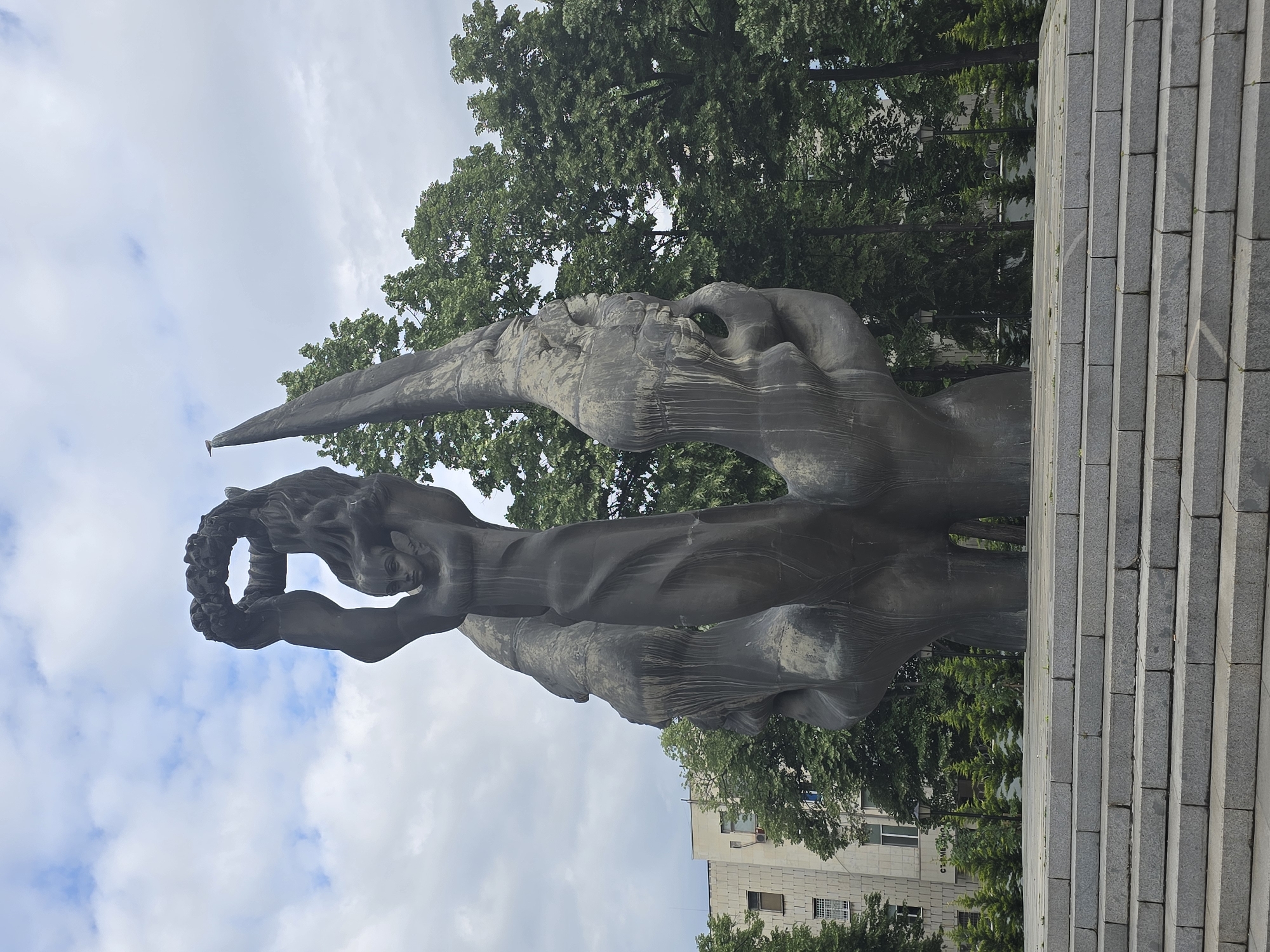The image depicts a monument with a dark-colored, abstract sculpture of a human figure holding a wreath overhead. The sculpture stands atop a set of gray stone steps. The background includes green trees and a cloudy sky. A building is visible in the distance. The statue is in the city of Unknown, Unknown.