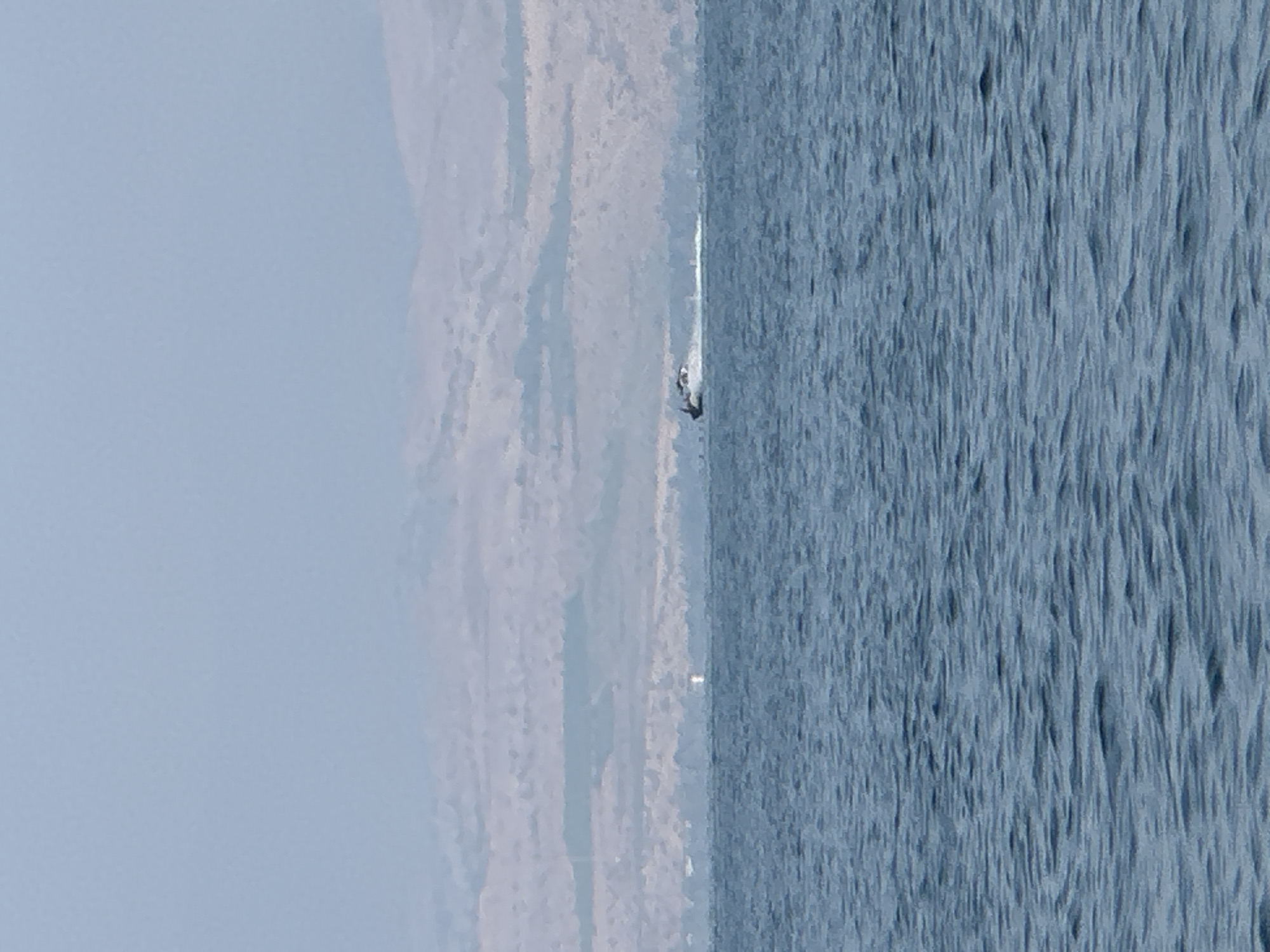 A wide shot captures the Sea of Galilee with a distant shoreline under a hazy sky. In the mid-ground, a dark-colored speedboat is actively moving across the water, creating a white wake behind it. The shoreline, appearing as a range of muted brown and beige hills, is largely obscured by atmospheric haze. A small, white marker buoy is visible in the water to the left of the speedboat. The water itself is a deep blue, with subtle ripples on its surface. The overall impression is of a serene, vast body of water with a singular object of human activity.