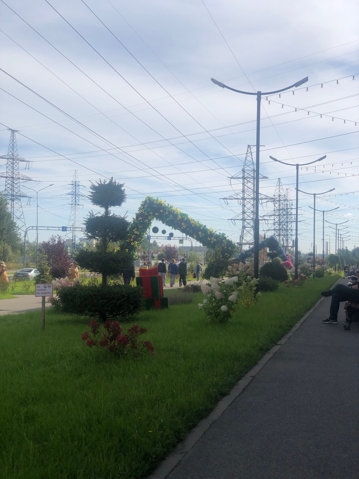 The image shows a park with a paved walkway on the right and a grassy area on the left. A decorative arch made of greenery and flowers spans across the walkway. In the grassy area, there are several manicured bushes, a large red and green gift box decoration, and a sign with Russian text that reads "ПО ГАЗОНУ НЕ ХОДИТЕ" (Do not walk on the lawn). Several people are walking in the background, and one person is sitting on a bench on the right side of the walkway. The scene is framed by numerous power lines and tall electrical towers, suggesting an urban or suburban environment. The sky is overcast. The location is identified as Pargolovo, Russia.