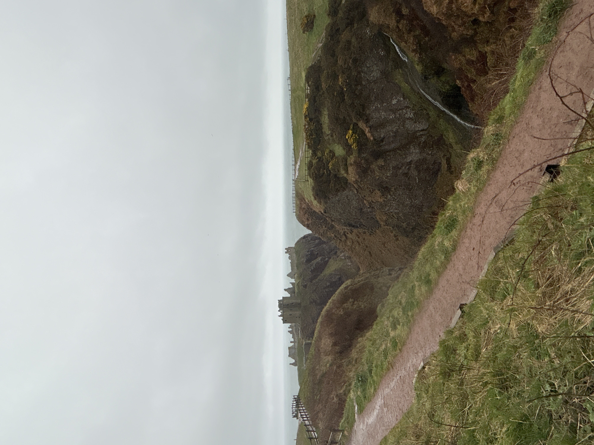 The image depicts the Dunnottar Castle in Stonehaven, United Kingdom. The castle ruins are situated on a rocky headland jutting into the North Sea. A narrow, muddy footpath runs along the edge of a cliff towards the castle. The sky is overcast and grey. A waterfall flows from the cliff edge. A wooden fence is visible along the path. The vegetation consists of grass and bushes, showing signs of dampness.