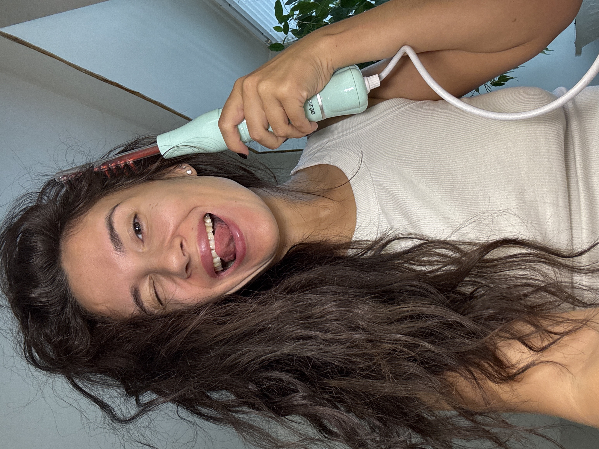 A young adult female, with long, dark, wavy hair, is depicted from the chest up in an indoor setting. She is wearing a white ribbed tank top. Her left eye is closed in a wink, and her mouth is open wide in a joyful expression, with her tongue slightly extended and teeth visible. A diamond stud earring is visible in her right ear. Her right hand holds a light mint green, handheld electronic device branded "GEZA" on its silver mid-section. The device features a translucent red comb-like attachment at its top, which appears illuminated, positioned near the woman's head and hair. A white electrical cord extends from the base of the device. The background consists of light blue walls, a visible white ceiling line in the upper right, a window with horizontal white blinds, and green foliage from an unseen plant on the right side of the frame. The specific location within the city of Chaikovsky, Russia, cannot be determined from the visual evidence provided in the image.