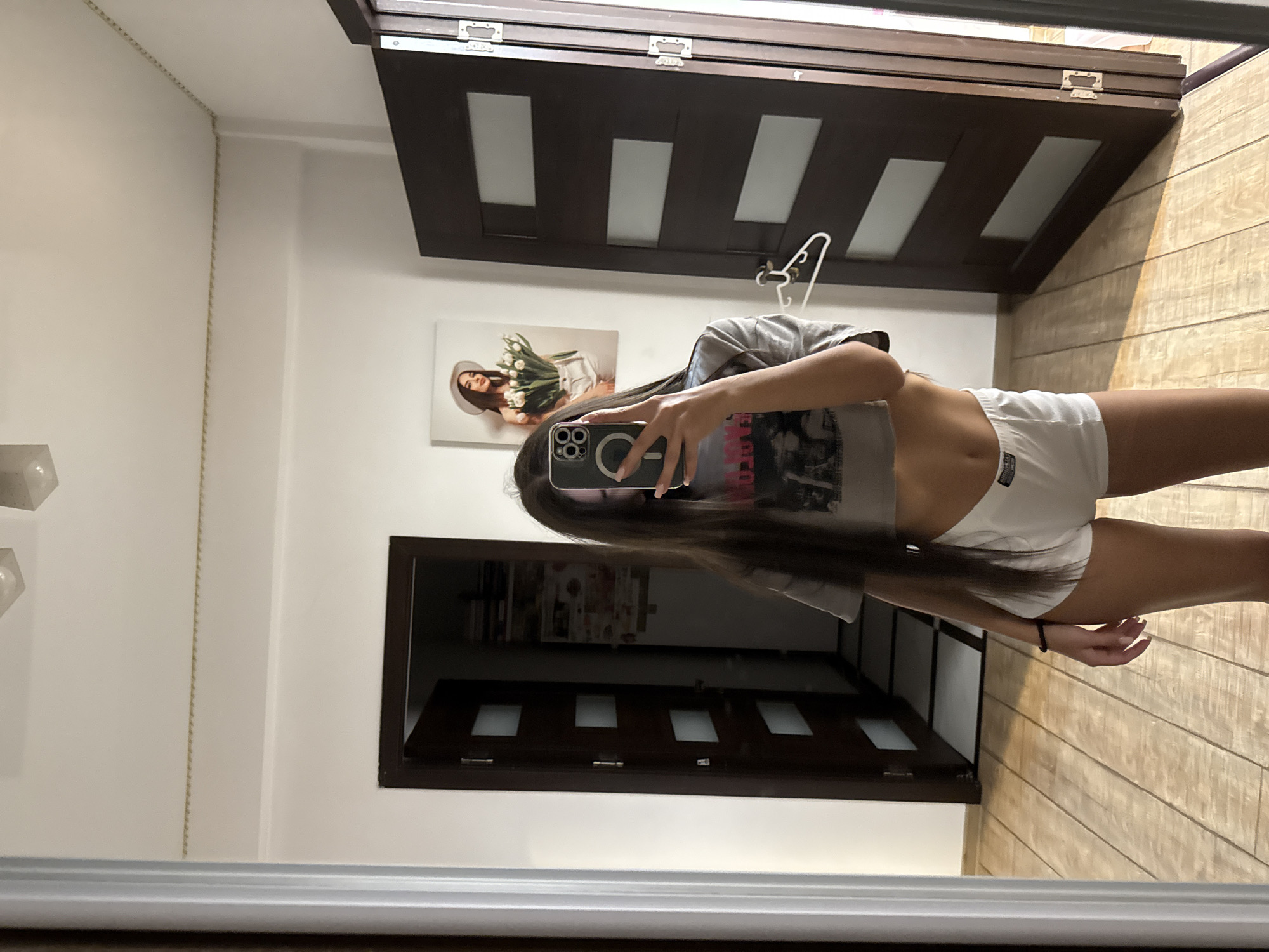 A young woman with long brown hair is taking a mirror selfie in a room. She is wearing a grey crop top and white shorts. She is holding a smartphone in her right hand. The room has wooden floors and two dark brown doors with frosted glass panels. There is a framed picture of a woman with tulips hanging on the wall. The background suggests an indoor setting, possibly a hallway or bedroom, within a residential building. The specific location within Chișinău, Moldova, cannot be determined from the visual information provided.