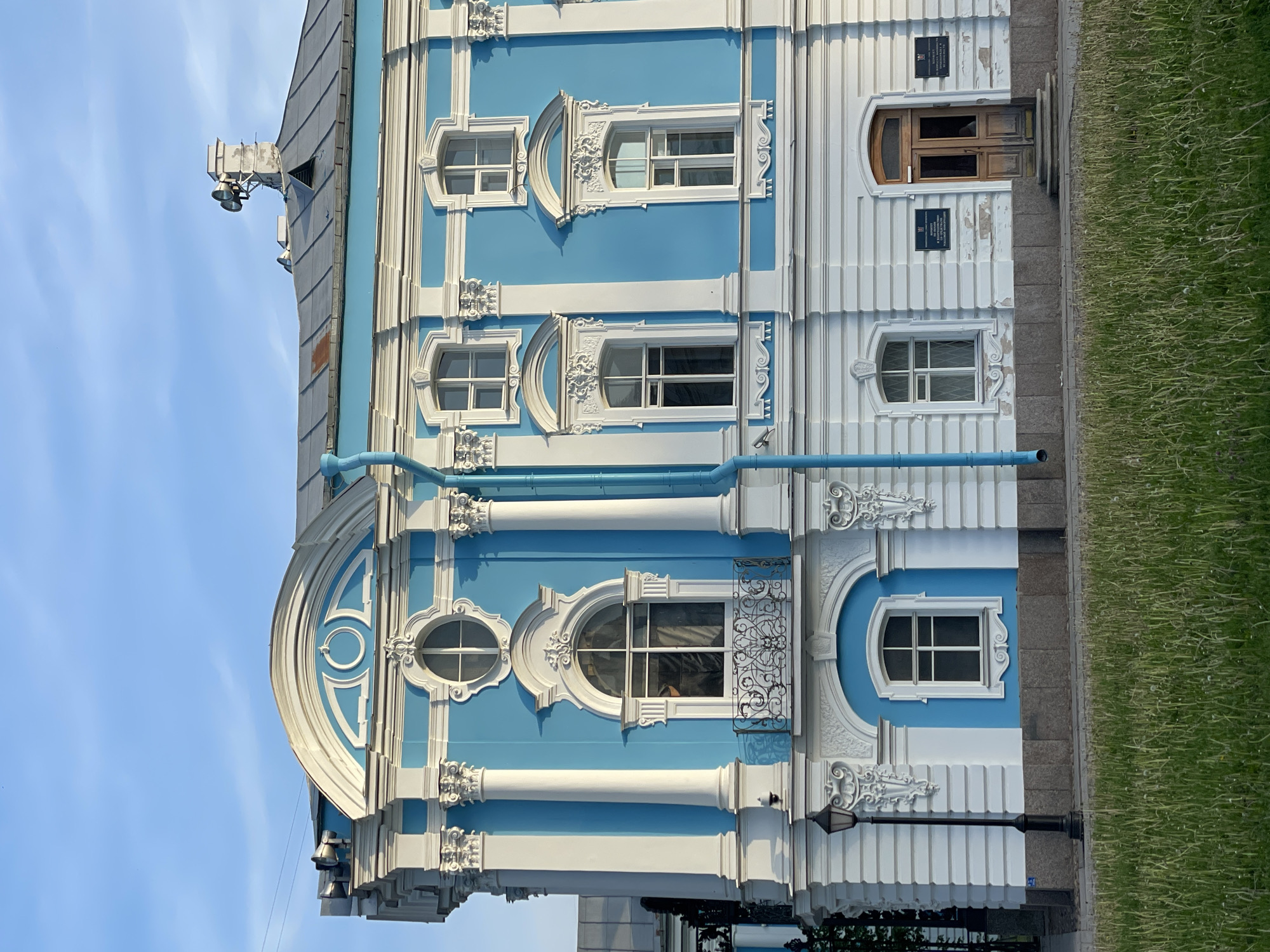 A partial exterior view of the Smolny Institute building in Saint Petersburg, Russia, is depicted under a clear, light blue sky with high sun illuminating the facade. The multi-story building features a light blue main wall accented with white Neoclassical architectural elements, including prominent columns with Corinthian capitals, cornices, and decorative window surrounds.
On the left side of the facade, a large arched window with a black wrought-iron balcony is centrally positioned, flanked by two white columns. Above this, an oval window is set within an ornate white frame, topped by a large semi-circular pediment decorated with a central circular motif. To the right of the pediment, a light blue downspout descends vertically along the facade.
Further right, the facade presents two levels of rectangular windows, each with white architraves and decorative elements. The roof, visible above the cornice line, is dark grey and metallic, featuring multiple vents and a prominent chimney-like structure supporting two horn-shaped loudspeakers mounted on a metal frame.
At ground level, the white base of the building exhibits horizontal banding and some areas of peeling paint. A traditional black lamppost stands on the far left. Towards the right, a recessed wooden double door with a small arched transom window serves as an entrance, accessed by two stone steps. Flanking the door are two dark blue plaques with white Russian text, indicating official institutions.
The immediate foreground consists of a strip of green grass, containing various blades and seed heads. Behind the grass, a dark grey granite curb or low wall separates it from a paved sidewalk area. No individuals or discernible actions are present in the scene.