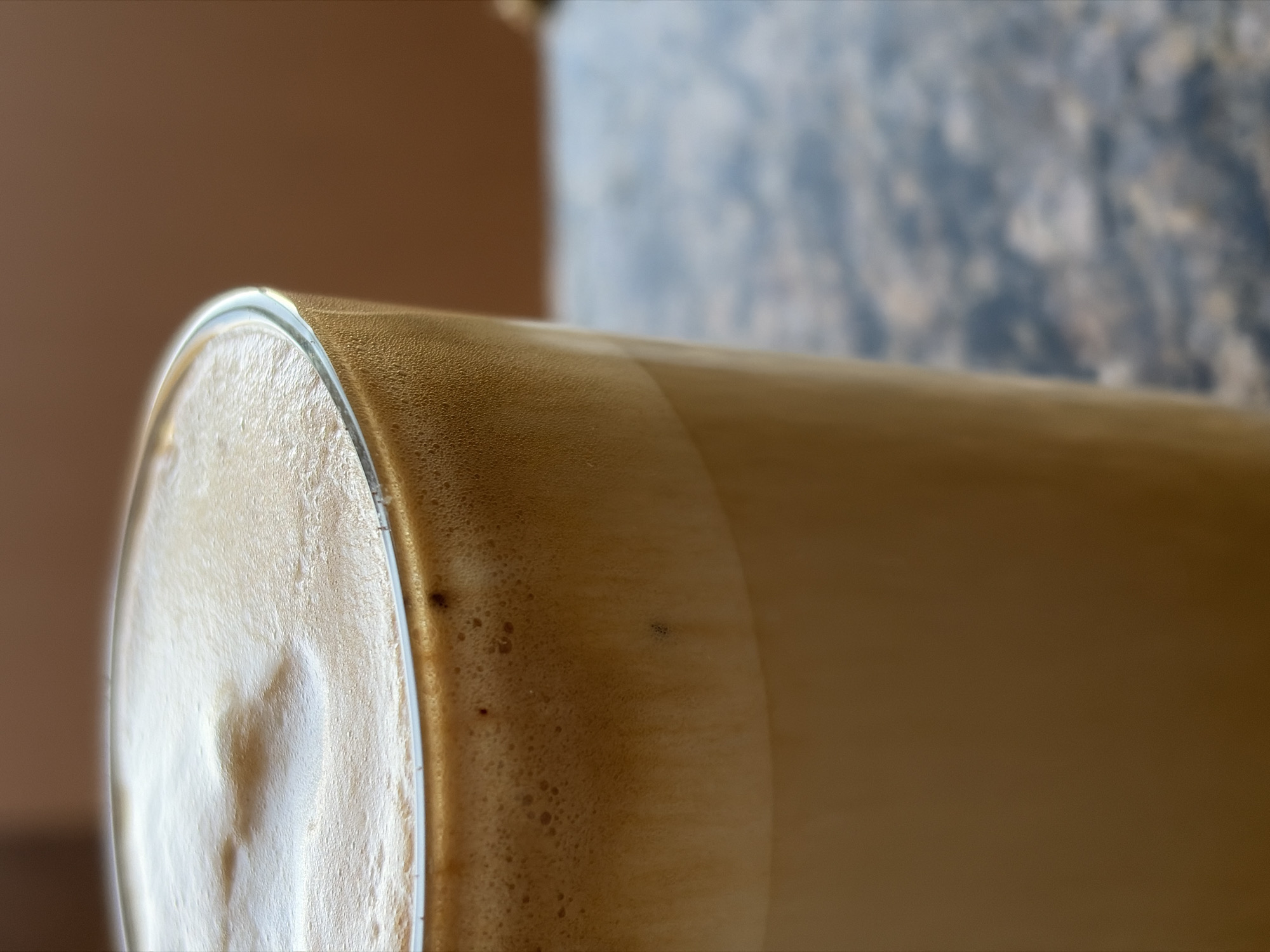 A close-up photograph shows a glass filled with a light brown, frothy beverage. The focus is on the top edge of the glass where the beverage meets the glass. The liquid is slightly blurred as it stretches away from the foreground. The background is out of focus and shows a beige wall and a mottled grey surface. The location is not identifiable as Sofia, Bulgaria, from the visual information present in the image.