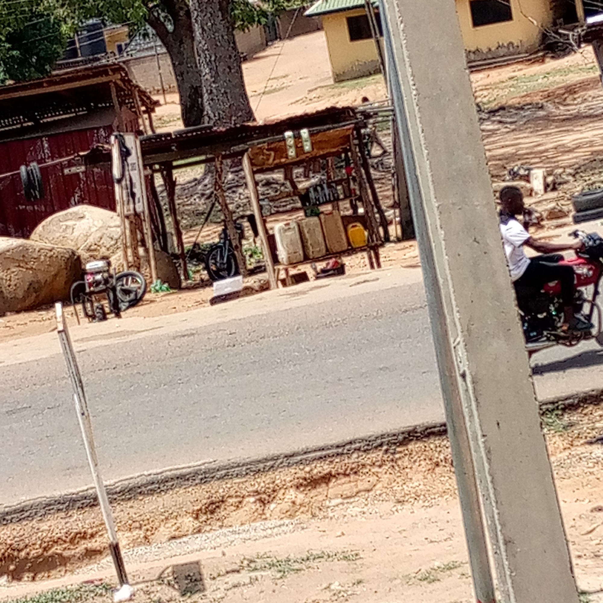 The image shows a street scene in Unknown, Unknown. A man rides a red motorcycle on the right side of the road. On the left, there is a small roadside structure with containers on display, possibly for sale. There are also some rocks and what appears to be a generator near the structure. In the background, there are trees and a building. A utility pole is in the foreground, slightly obstructing the view.