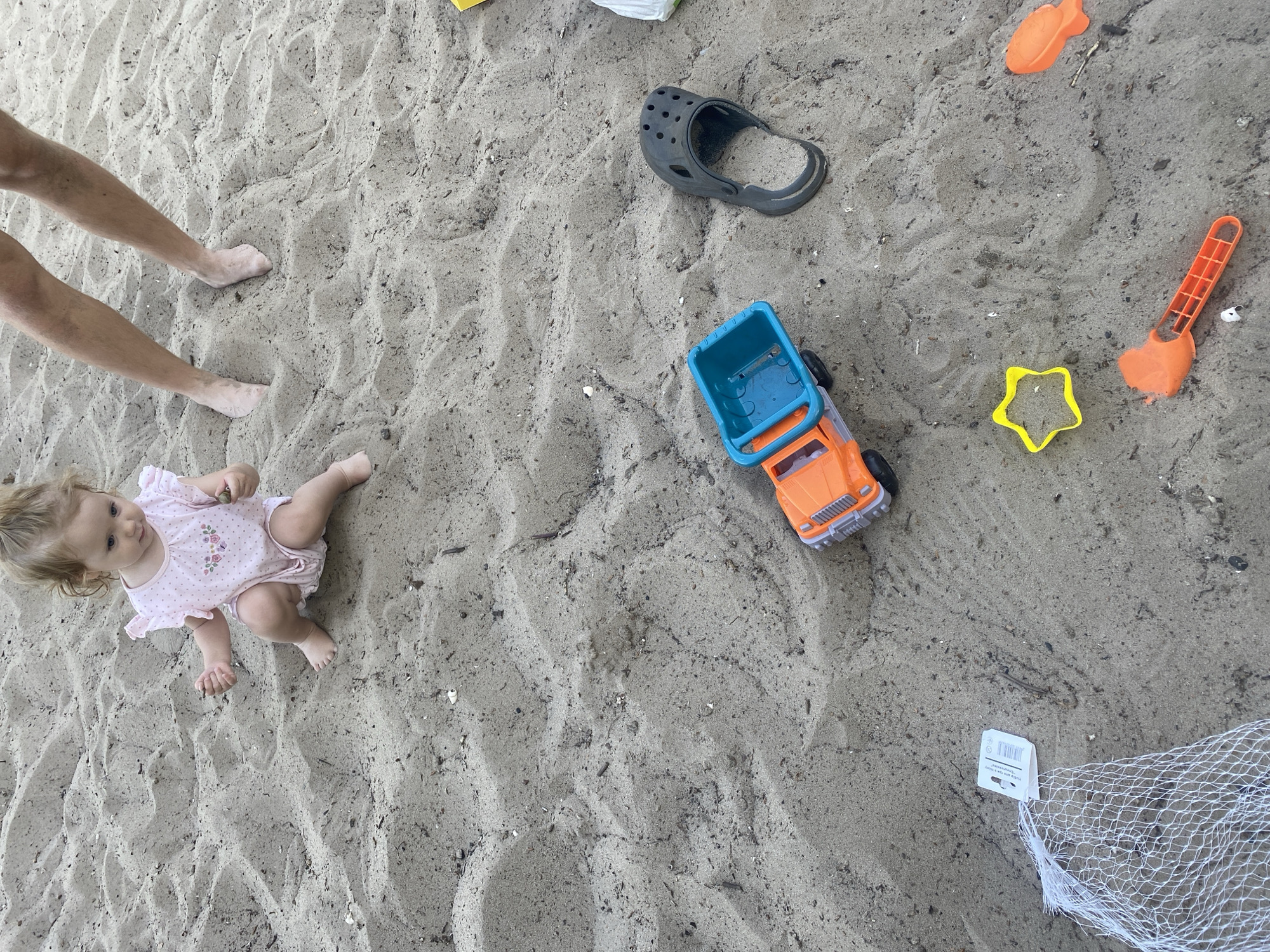 A blonde toddler wearing a pink romper sits on a sandy beach. She is holding two small pebbles in her hands. Her legs are bent and her feet are bare. To the right of the toddler, a pair of adult legs stand in the sand. In the foreground, several children's toys are scattered in the sand: an orange toy truck with a blue bucket, a black Croc sandal, a yellow star-shaped cookie cutter, an orange toy shovel, and an orange fish-shaped toy. A white mesh bag with a tag is visible to the left of the truck. The sand appears damp in some areas. The scene is set on a beach, possibly in Dnipro, Ukraine, given the context.