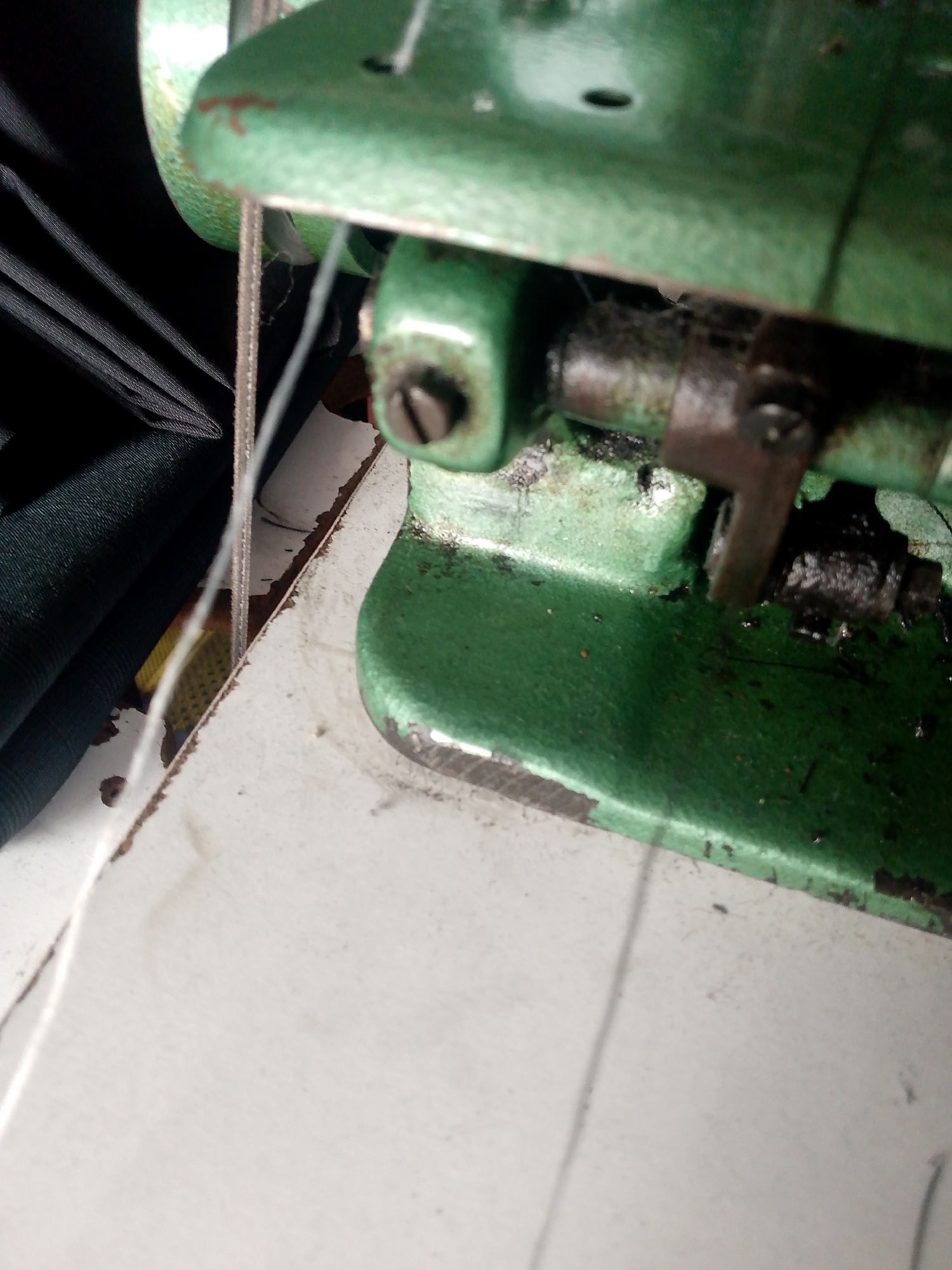 A close-up, low-angle shot shows a section of a green industrial sewing machine, likely situated on a white workbench, in Jauro Yinu, Nigeria. The machine appears aged, with areas of chipped paint revealing rust underneath. The focus is on the needle area and part of the presser foot mechanism, which is caked with dark grease and lint. A fine white thread is visible, running down from above and disappearing into the machine's workings. To the left, a stack of folded dark fabric, possibly black or dark green, is partially visible, suggesting the machine is in active use or recently used. The background is indistinct, but the white surface beneath the machine appears worn and marked. The lighting is somewhat dim, casting soft shadows and highlighting the texture of the fabric and the machine's surface. There are no people or animals visible in the frame.