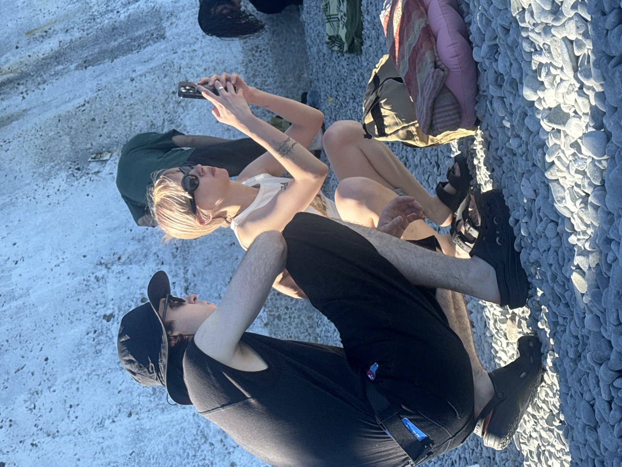 Two individuals are crouched on a pebbled surface, likely a beach, in the city of Partenit, Ukraine. The person on the left wears a black bucket hat, sunglasses, a black sleeveless shirt, and black pants, and has a blue tag attached to their belt loop. They are also wearing black Crocs. The person on the right has short, blonde hair, wears sunglasses, and a white tank top with a tattoo on their arm. They are holding a smartphone in their right hand, appearing to take a picture or video. In the background, another person in a green shirt is partially visible, also crouching. Various items, including bags and blankets, are scattered on the ground around them. The environment appears to be outdoors with a large, textured concrete or stone wall in the background. The lighting suggests it is daytime.