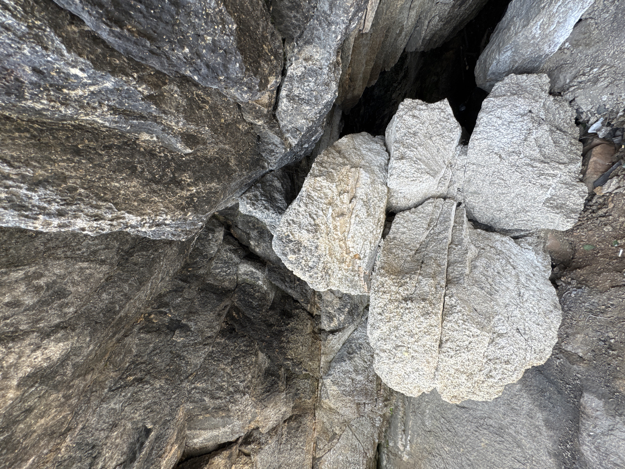 This is a close-up, eye-level, outdoor, medium shot of a pile of weathered, light gray boulders stacked in the foreground, nestled against a dark gray, rocky overhang that recedes into shadow. The boulders are rough and granular in texture, with visible cracks and indentations. The rock formation is substantial, with large, angular segments creating a sense of depth and cavernous space. The environment suggests a natural, possibly coastal or mountainous, setting, with some dry dirt and debris visible at the base of the rocks. The lighting is natural, creating distinct shadows and highlighting the varied textures of the stone. There are no people or animals visible in the image. This scene is located at Dom Otdykha Zolotoy Plyazh, Russia.