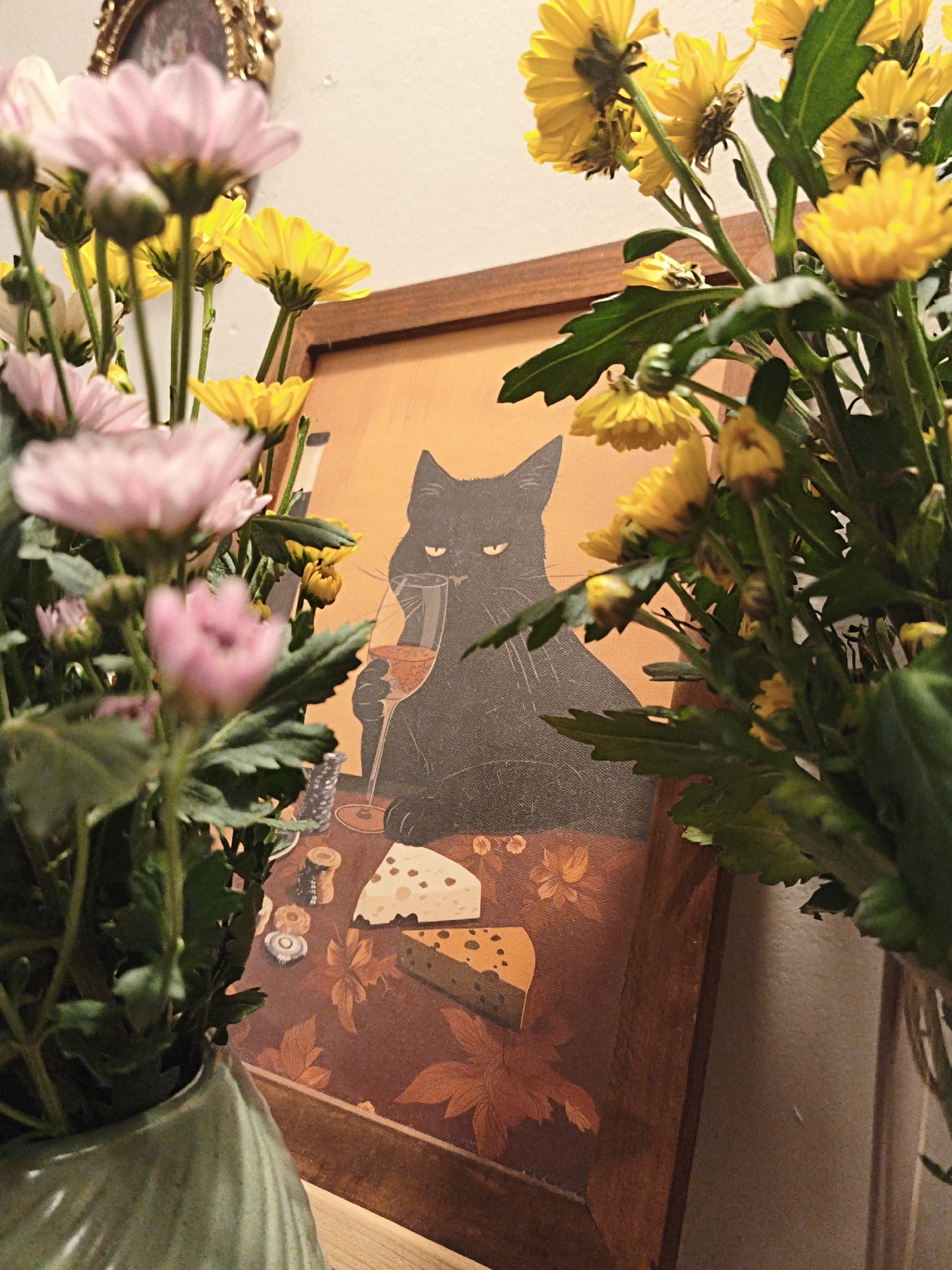 A framed painting of a black cat holding a glass of red wine and seated at a table with cheese and what appears to be spools of thread is partially obscured by bouquets of pink and yellow daisies. The painting is positioned on a wooden surface, possibly a shelf or table, and is framed by a dark wooden frame. The background is a plain, light-colored wall. The scene is illuminated by warm, artificial light, casting soft shadows. The overall impression is one of a cozy, slightly whimsical indoor setting. The location in the city of Bac Nha Trang Ward, Vietnam, is not discernible from the image.