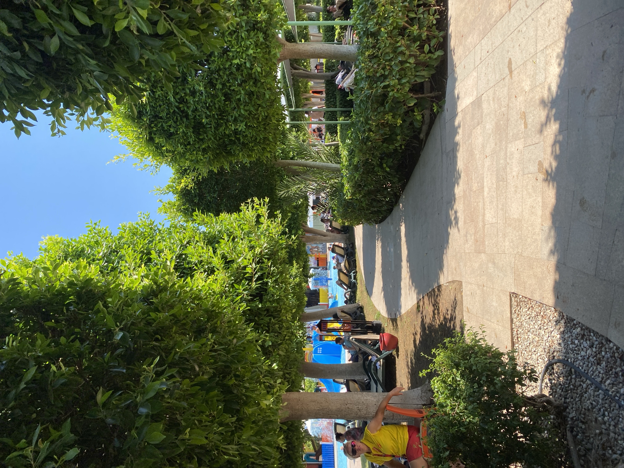 The image shows a sunlit outdoor area, likely a resort or water park, in Aksu, Turkey. Tall, manicured trees line a paved pathway that curves to the right. On the left, a person wearing a yellow shirt and red shorts, possibly a lifeguard, sits on an orange chair under a tree. Behind them, lounge chairs are set up on a grassy area, with people relaxing. In the background, a bright blue water slide and splash pool are visible, indicating recreational facilities. Lush green bushes form a dense hedge along the right side of the pathway. The sky is clear and blue.